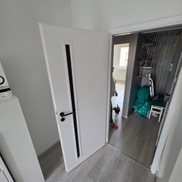 BART-BUD BARTOSZ ŁOŚ WYKOŃCZENIA WNĘTRZ - Nowe białe drzwi wewnętrzne z czarną klamką i pionowym panelem, zamontowane w jasnym korytarzu z szarymi panelami podłogowymi. W tle widać fragment pokoju z oknem i osobę z małym psem.