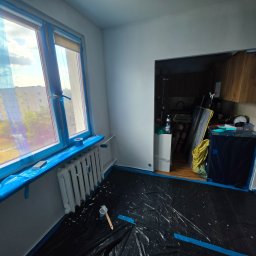 BART-BUD BARTOSZ ŁOŚ WYKOŃCZENIA WNĘTRZ - Pokój w trakcie remontu, z oknami i grzejnikiem precyzyjnie zabezpieczonymi niebieską folią. Podłoga pokryta czarną plandeką, na której leży pędzel i puszka farby. Widoczne przejście do kuchni.
