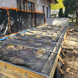 BART-BUD BARTOSZ ŁOŚ WYKOŃCZENIA WNĘTRZ - Przygotowanie do wylewania betonowej płyty tarasu przy białym domu, z drewnianym szalunkiem, zbrojeniem i małymi kopcami betonu na czarnej folii. Ściana domu zabezpieczona czarną folią...