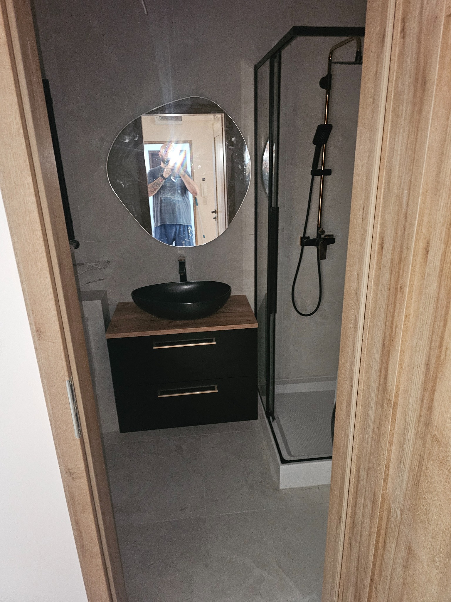 Nowoczesna łazienka z kabiną prysznicową, umywalką nablatową na szafce z szufladami i lustrem o nieregularnym kształcie. Widok zza otwartych drzwi.