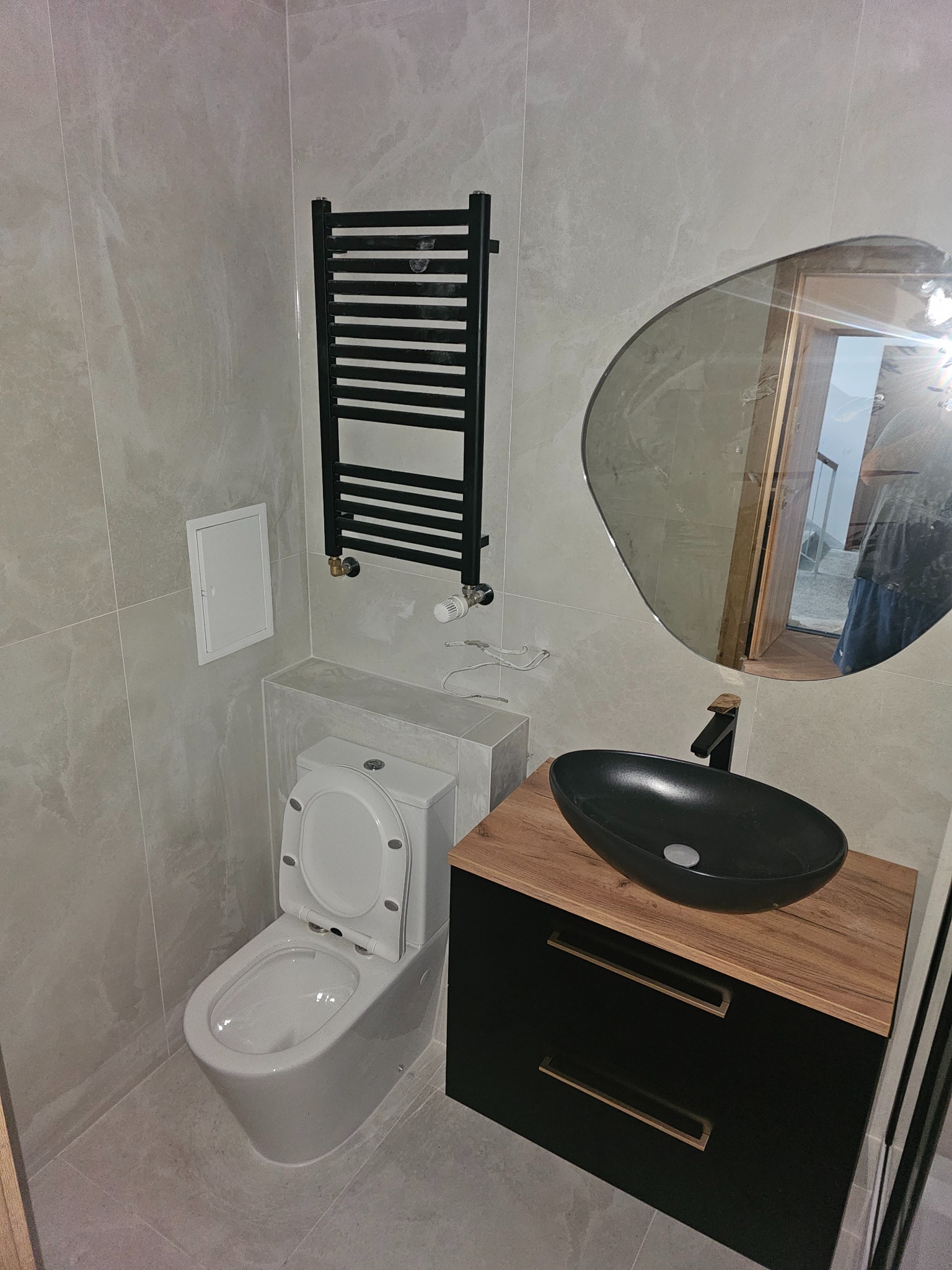 Nowoczesna łazienka z umywalką nablatową, grzejnikiem drabinkowym i toaletą. Szare płytki imitujące kamień. Nietypowe lustro dodaje charakteru.