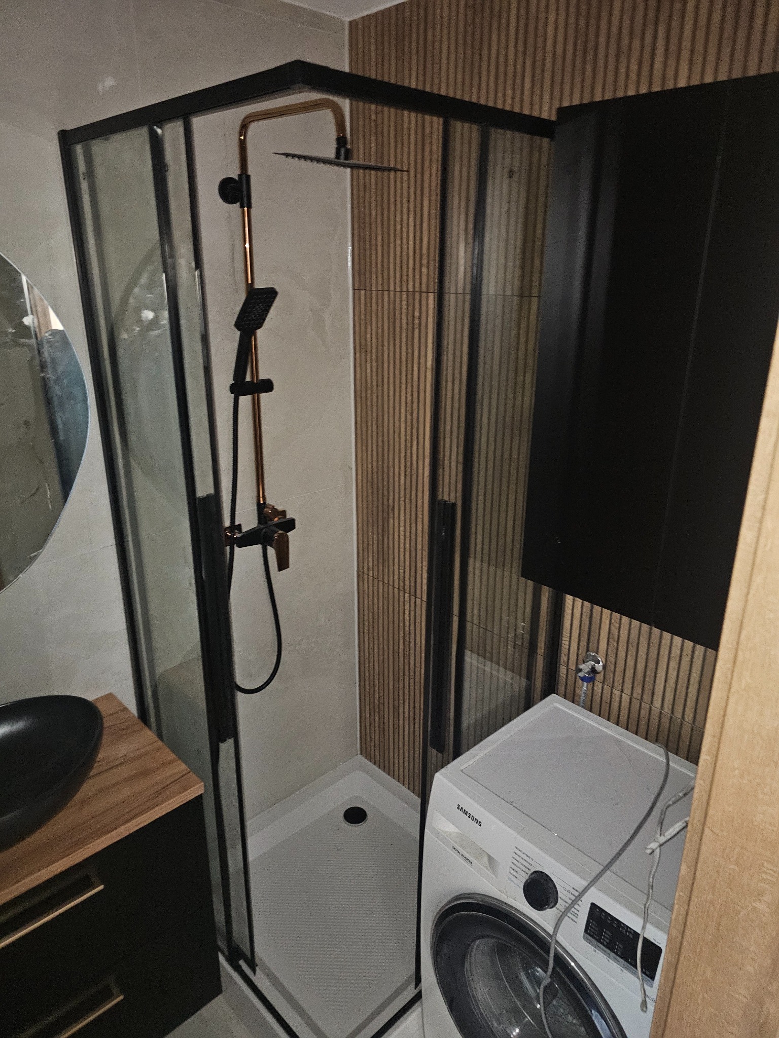 Nowoczesna kabina prysznicowa z czarną armaturą i deszczownicą, umywalka z szafką i pralka w wykończonej łazience z drewnopodobnymi panelami ściennymi.