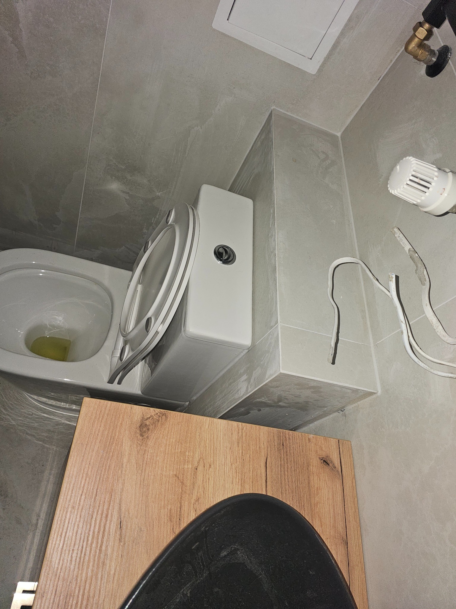 Szara łazienka w trakcie montażu toalety. Widoczne płytki, fragment szafki i wystające kable. Ujęcie z góry, podkreślające etap prac wykończeniowych.