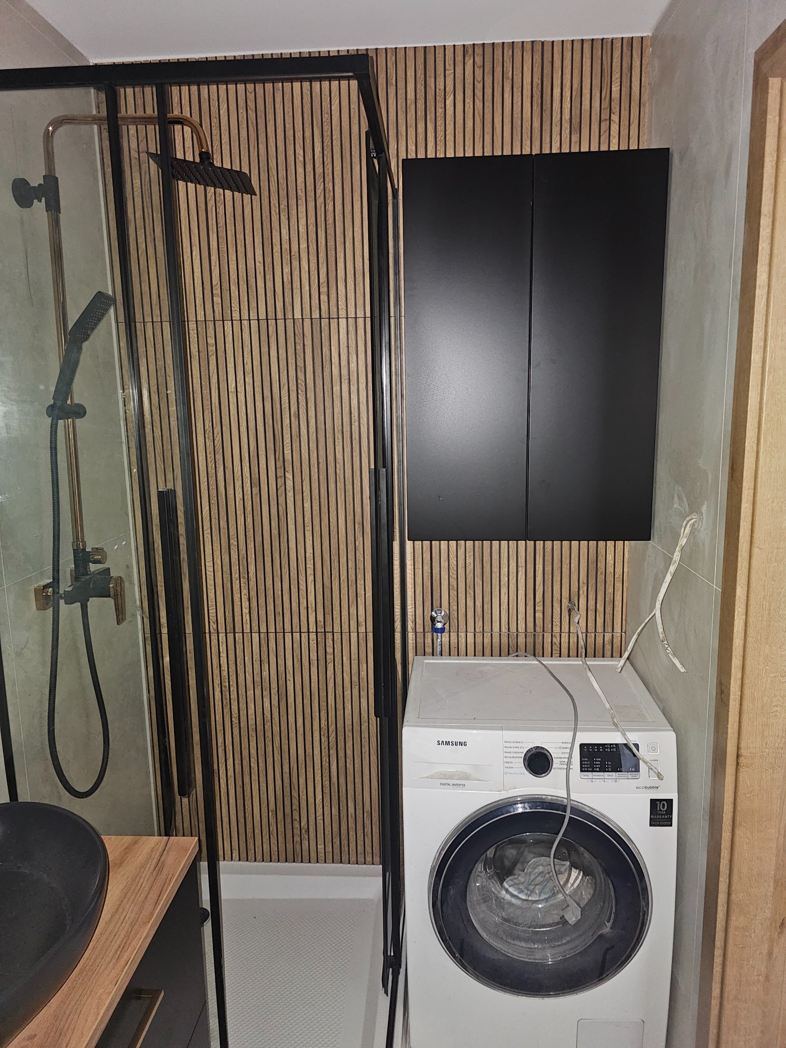 Nowoczesna łazienka po remoncie: kabina prysznicowa z czarnymi ramami, drewniana ściana z lamelami i pralka Samsung pod szafką. Minimalistyczny design.