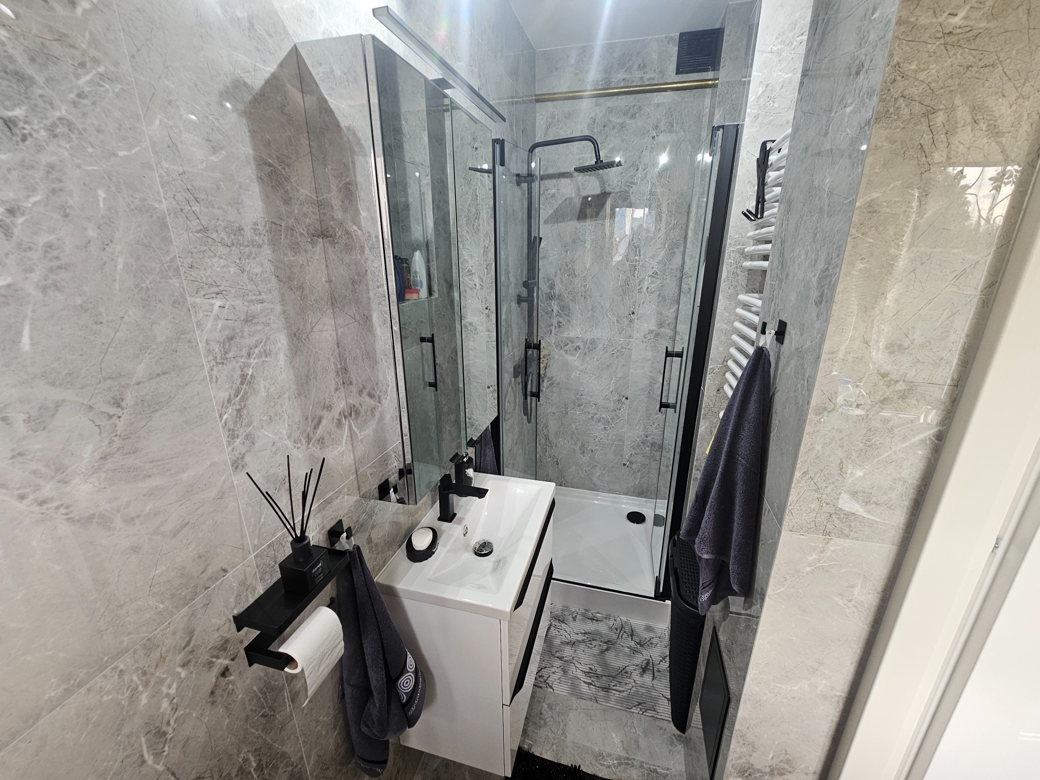 Elegancka, szara łazienka po remoncie: kabina prysznicowa typu walk-in z czarną armaturą, umywalka z szafką i marmurowe płytki na ścianach. Minimalistyczny design.
