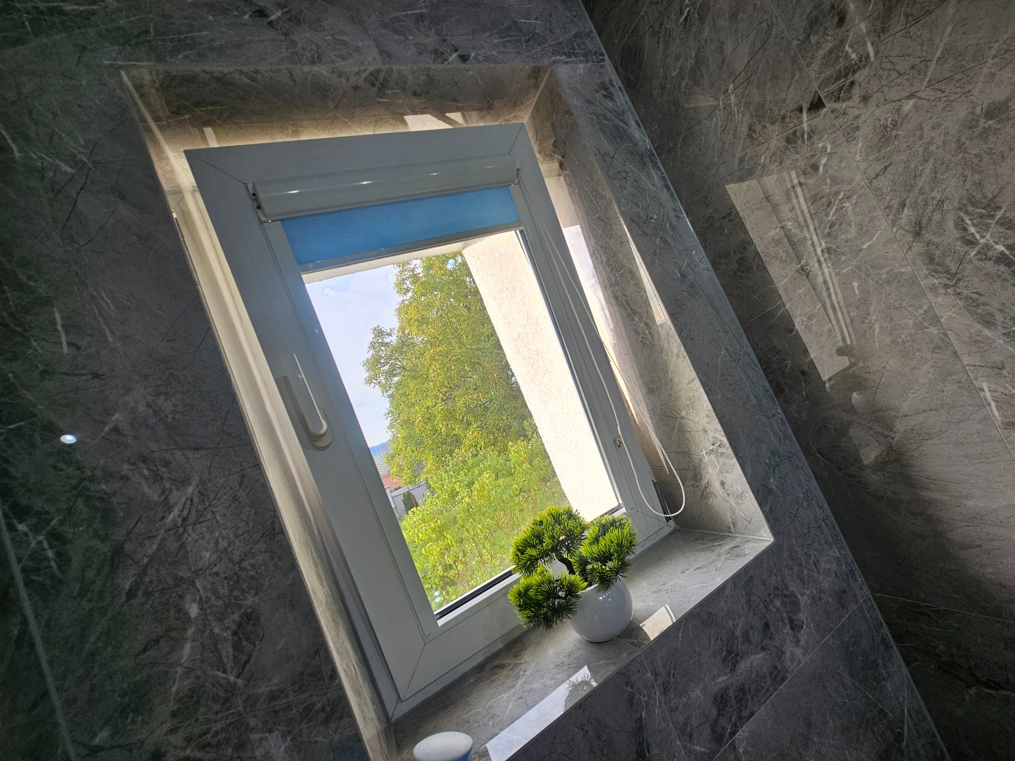 Wykończona ściana i parapet z szarego marmuru w łazience, okno z roletą i dekoracyjną rośliną w doniczce. Widok na zieleń za oknem.