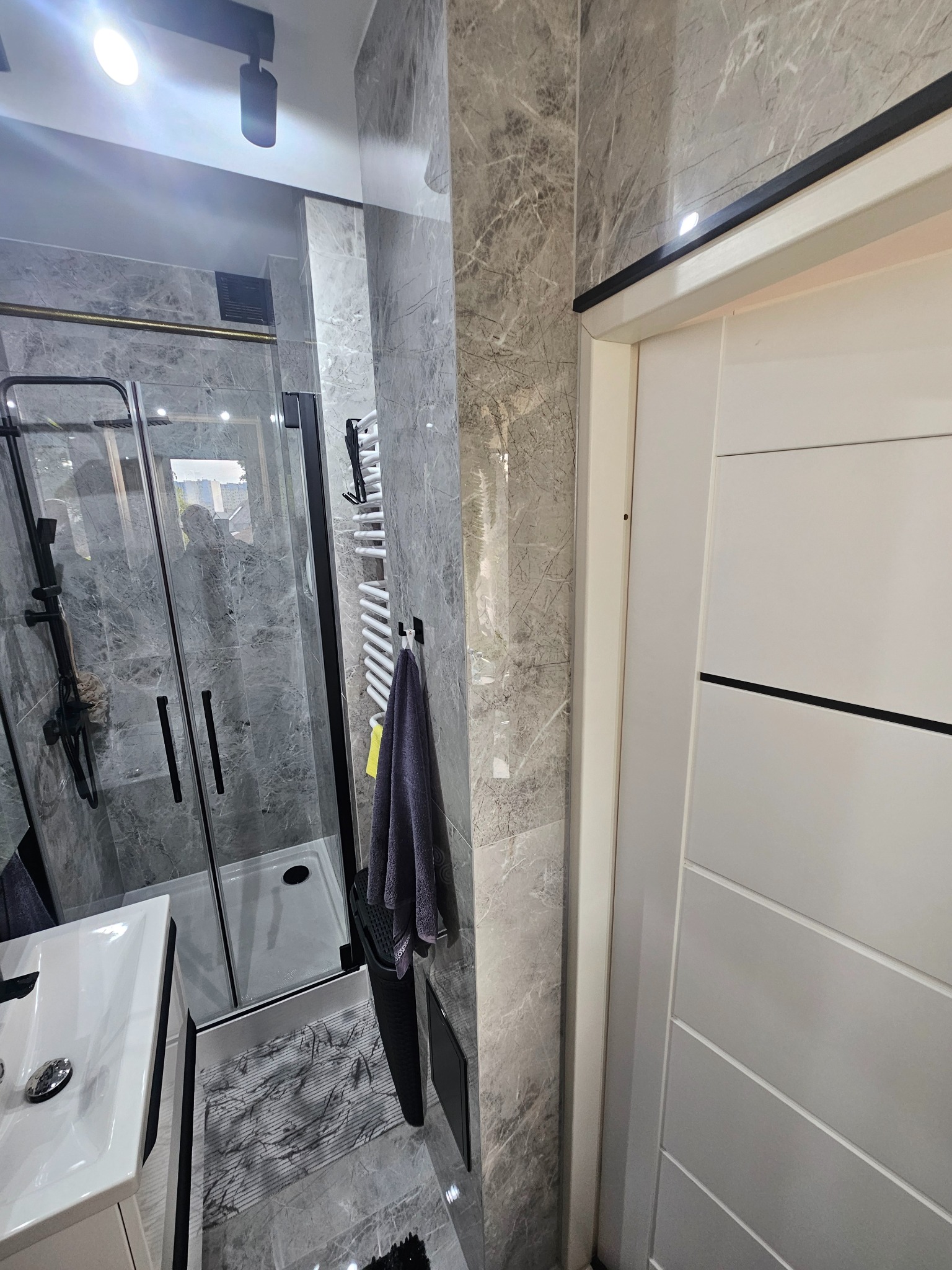 Nowoczesna łazienka z kabiną prysznicową typu walk-in, marmurowymi płytkami ściennymi, grzejnikiem drabinkowym i białą umywalką. Minimalistyczny design.