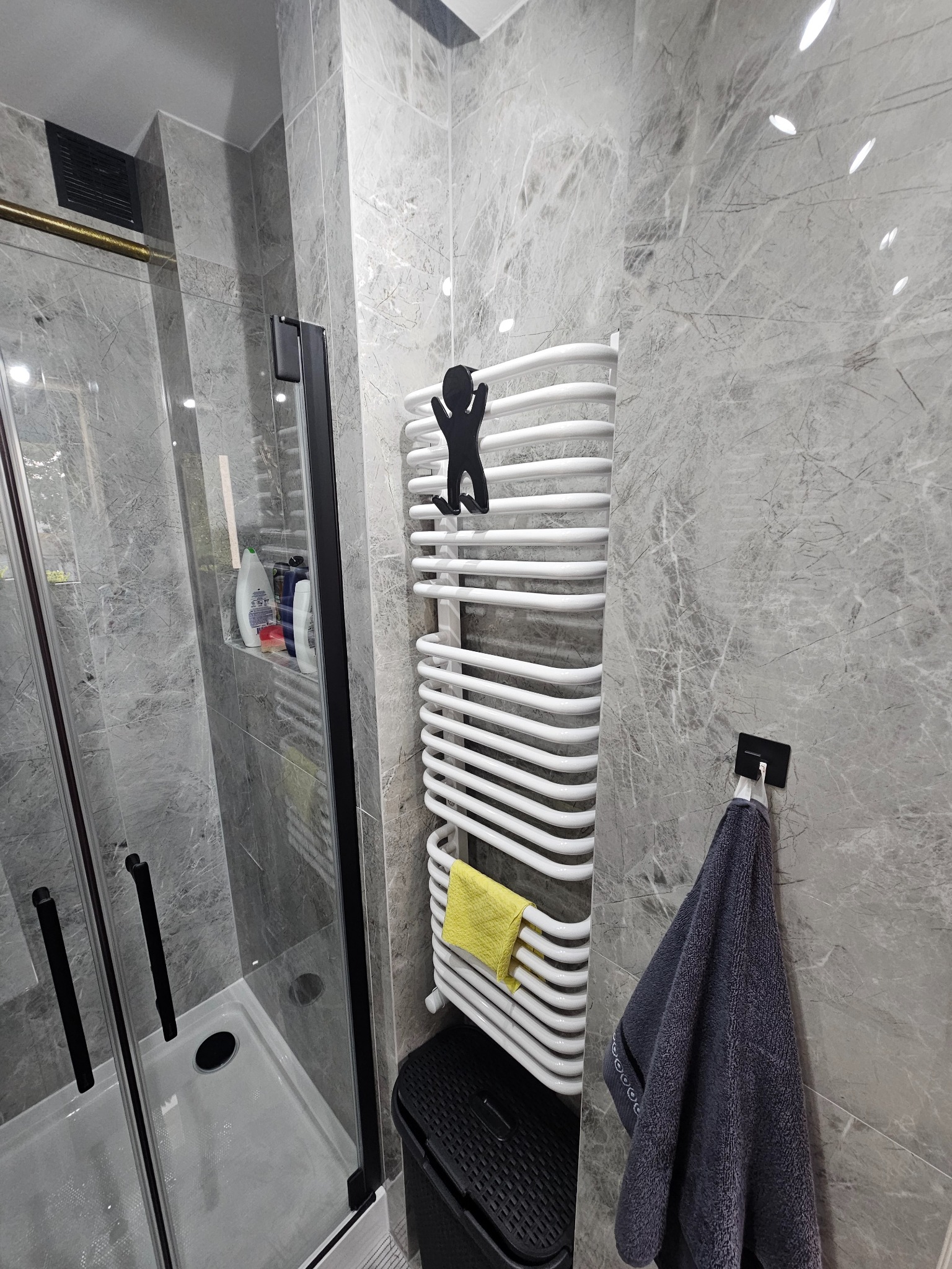 Wykończona łazienka z kabiną prysznicową, grzejnikiem drabinkowym z ozdobnym wieszakiem i marmurowymi płytkami ściennymi w odcieniach szarości.