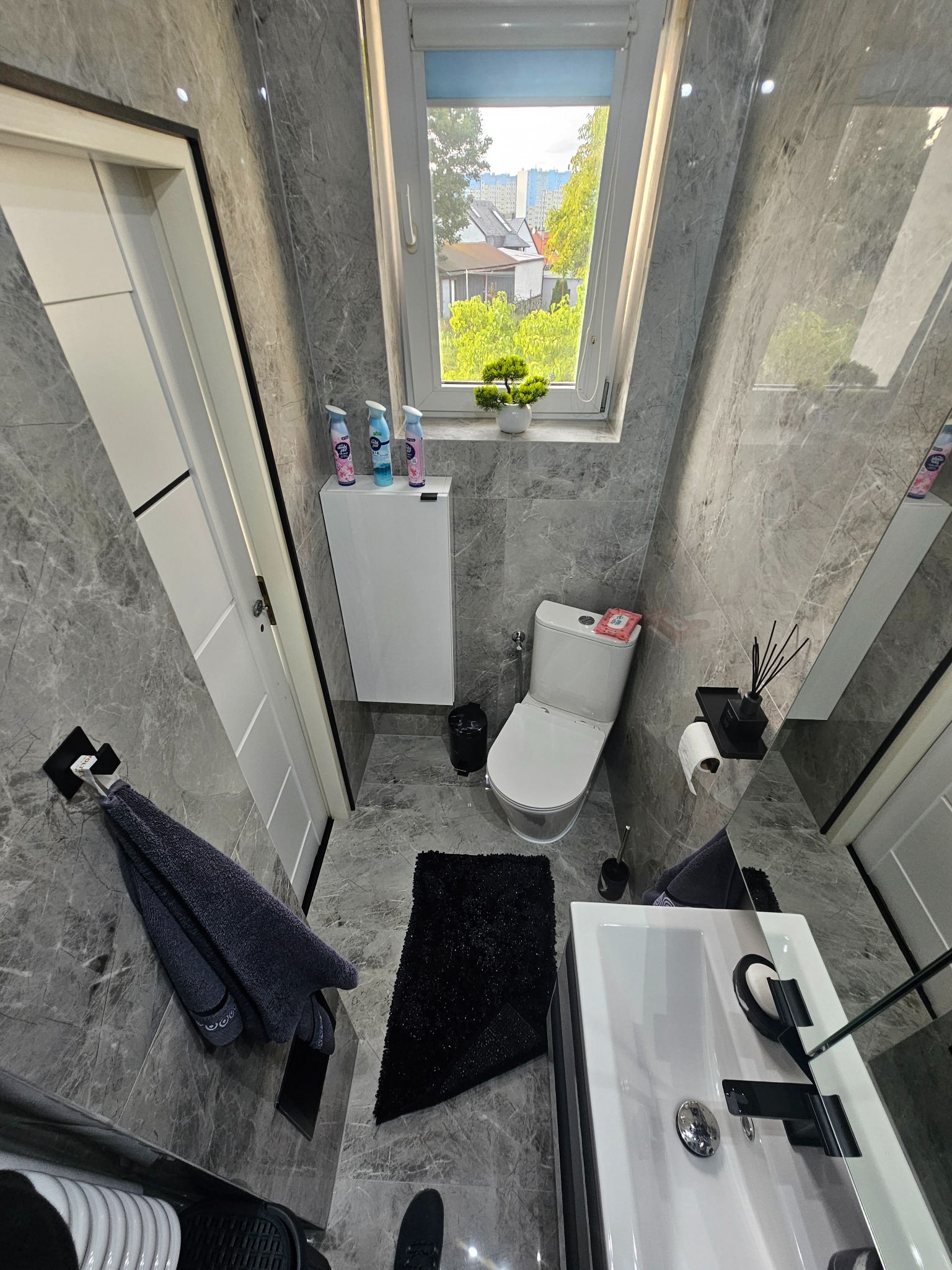 Nowoczesna łazienka z marmurowymi płytkami, białym wyposażeniem i oknem dachowym z widokiem na zieleń. Czarny dywanik i ręcznik dodają kontrastu.