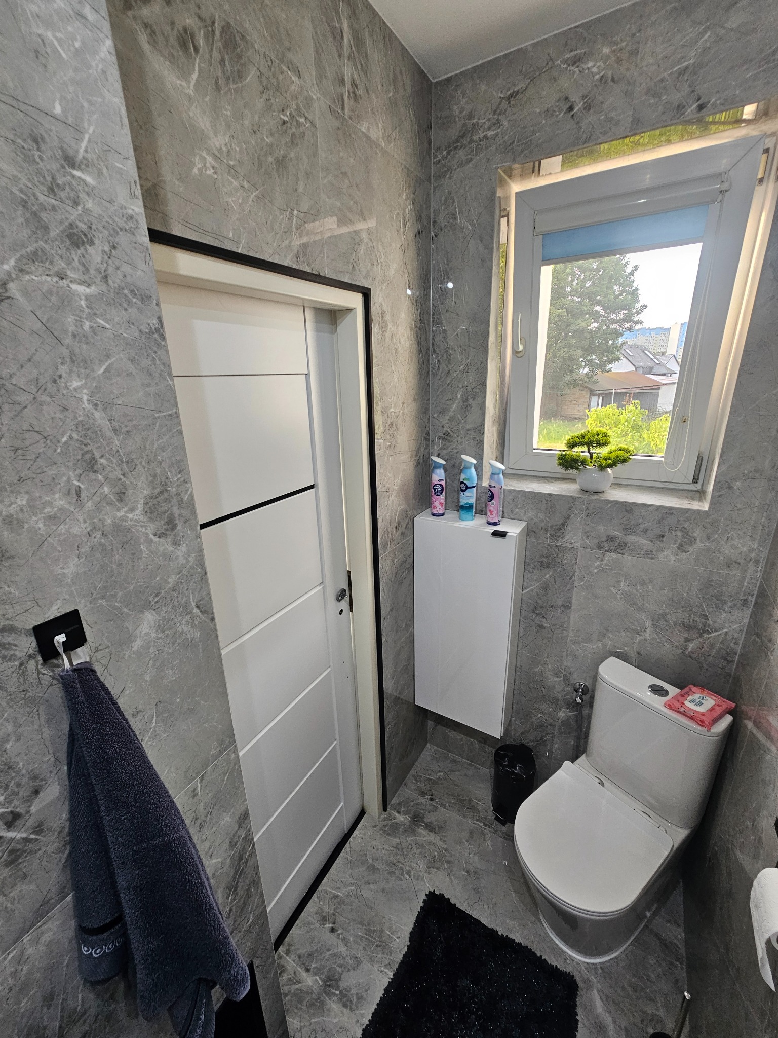 Nowoczesna łazienka w szarościach z marmurowymi płytkami. Biała szafka, toaleta i drzwi. Roślina w oknie dodaje naturalnego akcentu.