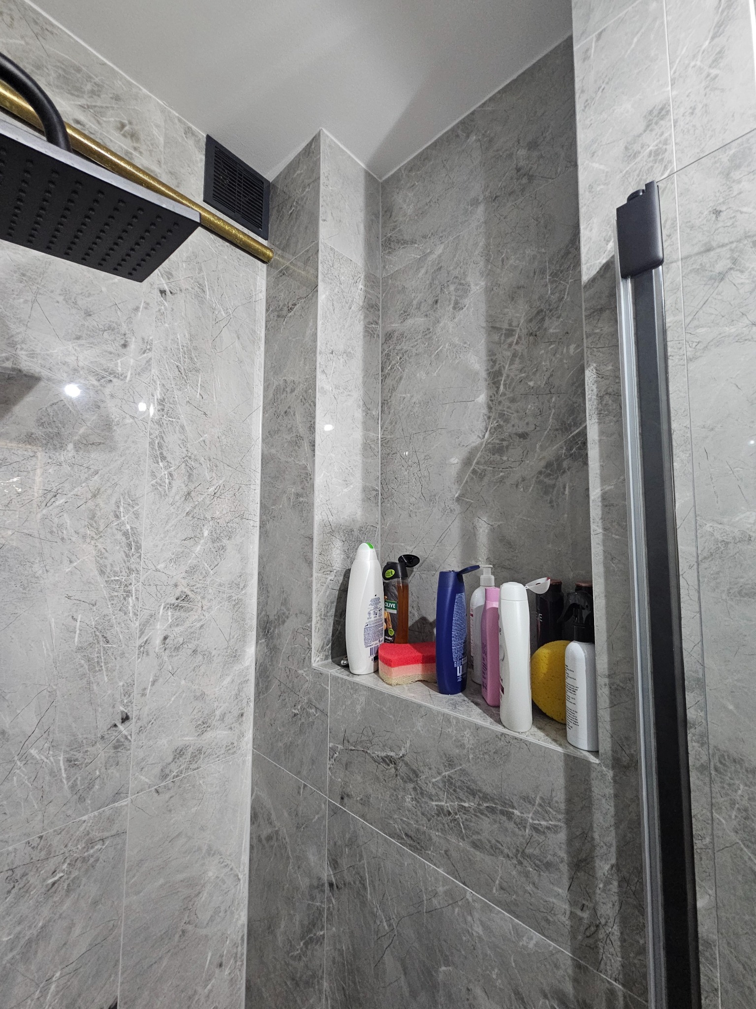 Nowoczesna kabina prysznicowa wyłożona szarymi płytkami imitującymi marmur. Widoczna czarna deszczownica z złotym ramieniem i wnęka z kosmetykami.