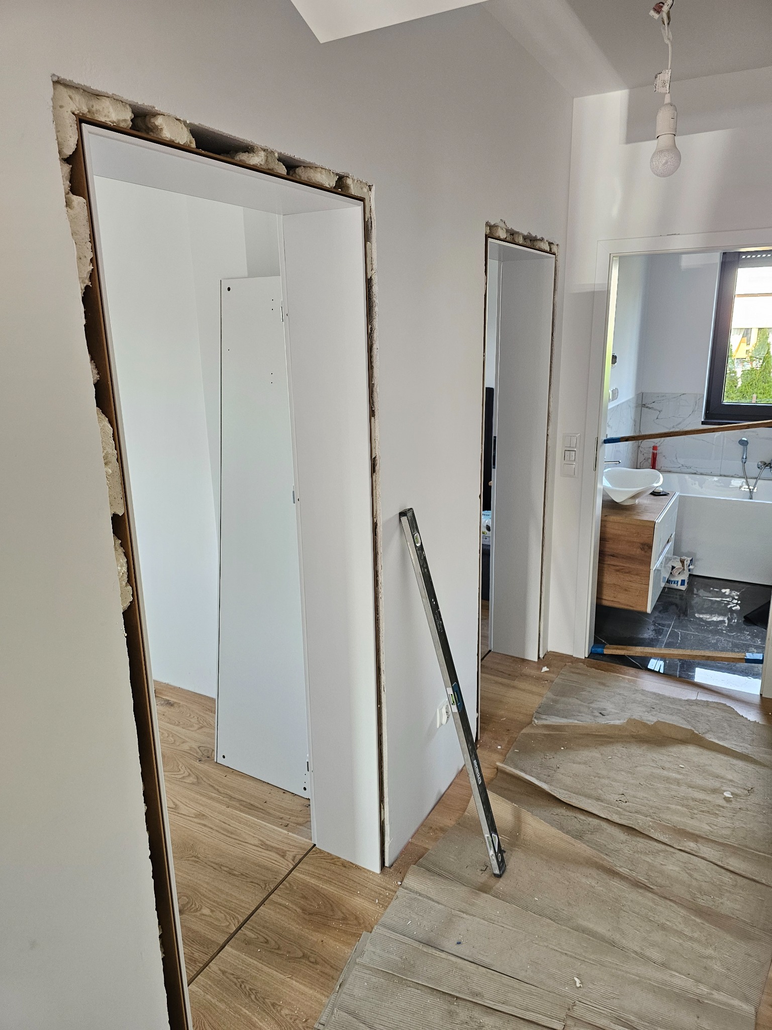 Montaż ościeżnic w nowym mieszkaniu. Widoczne surowe otwory drzwiowe, częściowo wypełnione pianką montażową. Poziomica oparta o ścianę. W tle widoczna łazienka.