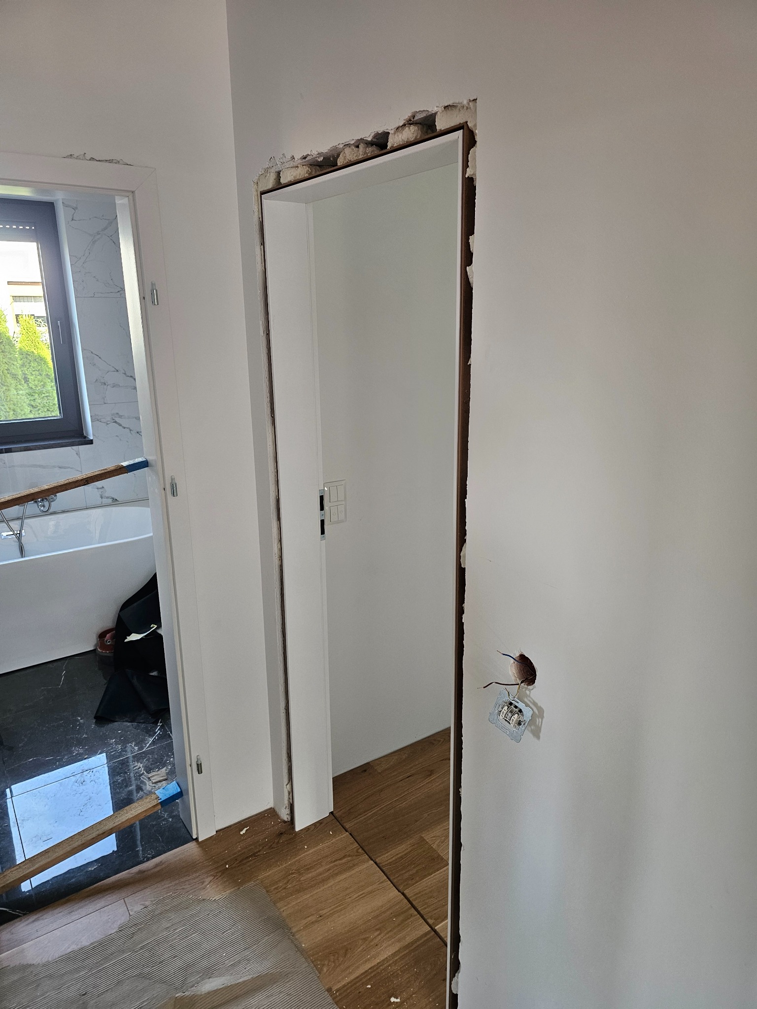 Wnętrze w trakcie remontu: widoczne futryny drzwiowe, fragment wykończonej podłogi i przygotowane ściany. W tle łazienka z oknem.