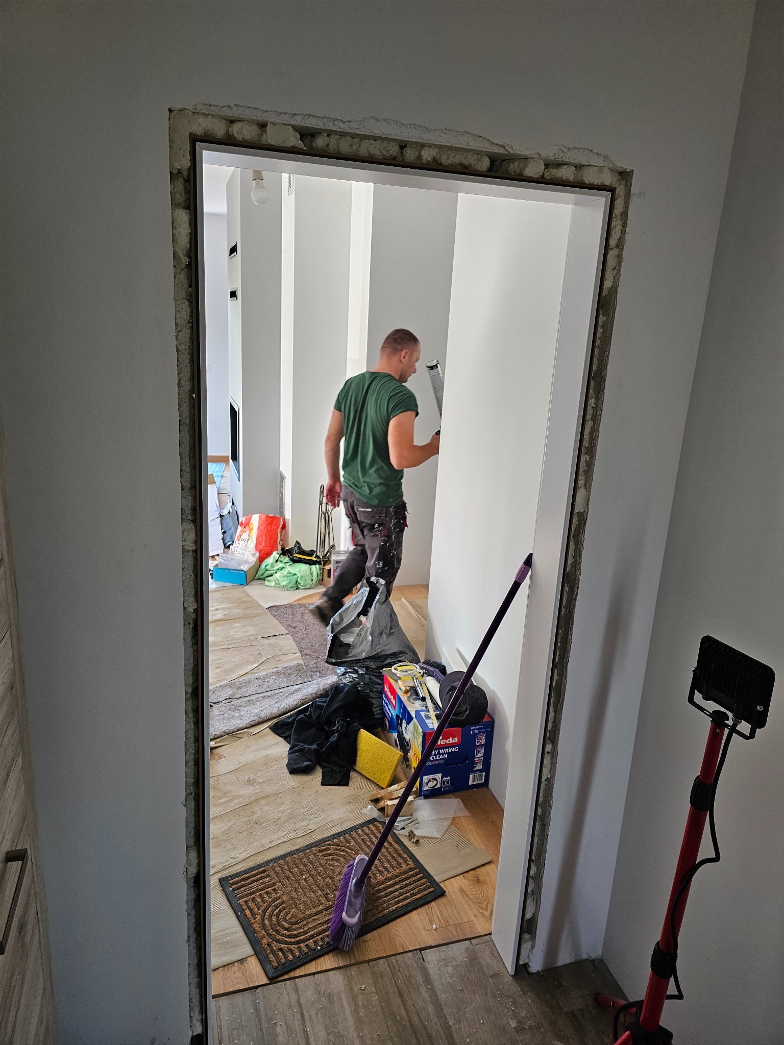 Montaż ościeżnicy drzwiowej: mężczyzna uszczelnia ramę silikonem, widoczne narzędzia i fragment wnętrza w trakcie prac wykończeniowych, oświetlenie robocze.