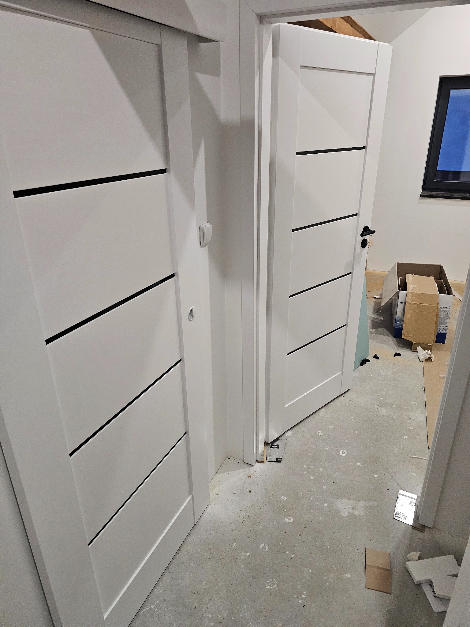 Dwa białe, nowoczesne drzwi z czarnymi, poziomymi wstawkami. Jeden zestaw drzwi jest zamontowany, a drugi otwarty, ukazując surowy stan podłogi i fragment okna.