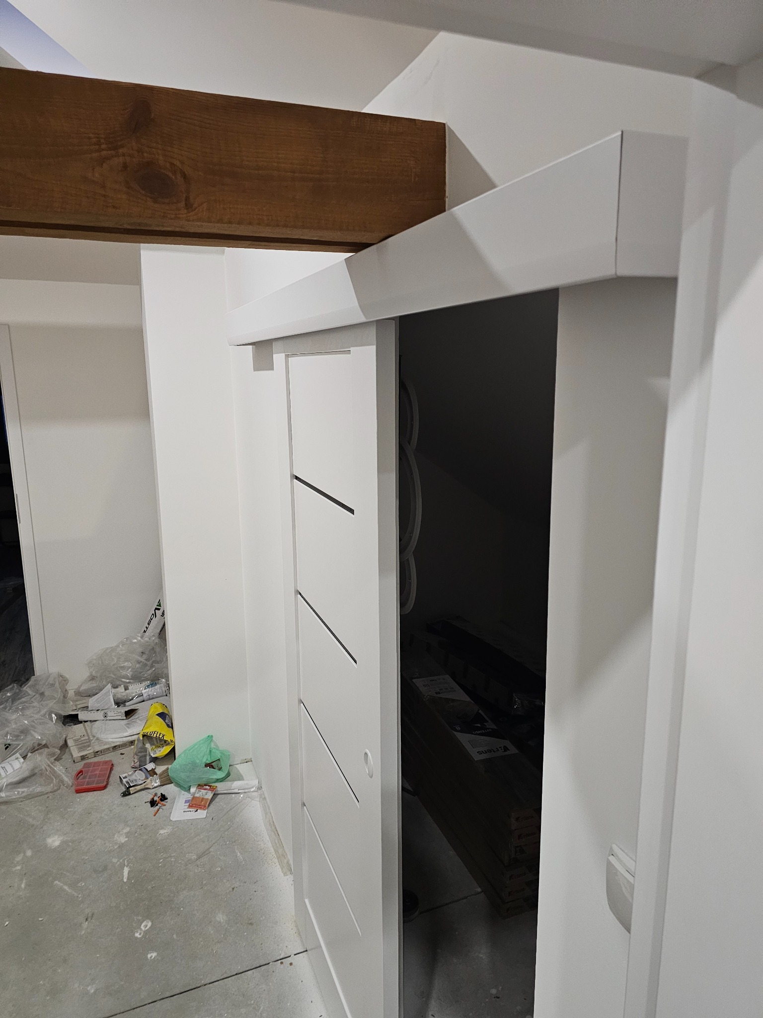 Białe, przesuwne drzwi z trzema czarnymi paskami, zamontowane w nowym, wykończonym wnętrzu. W tle widoczna drewniana belka, a na podłodze materiały budowlane.