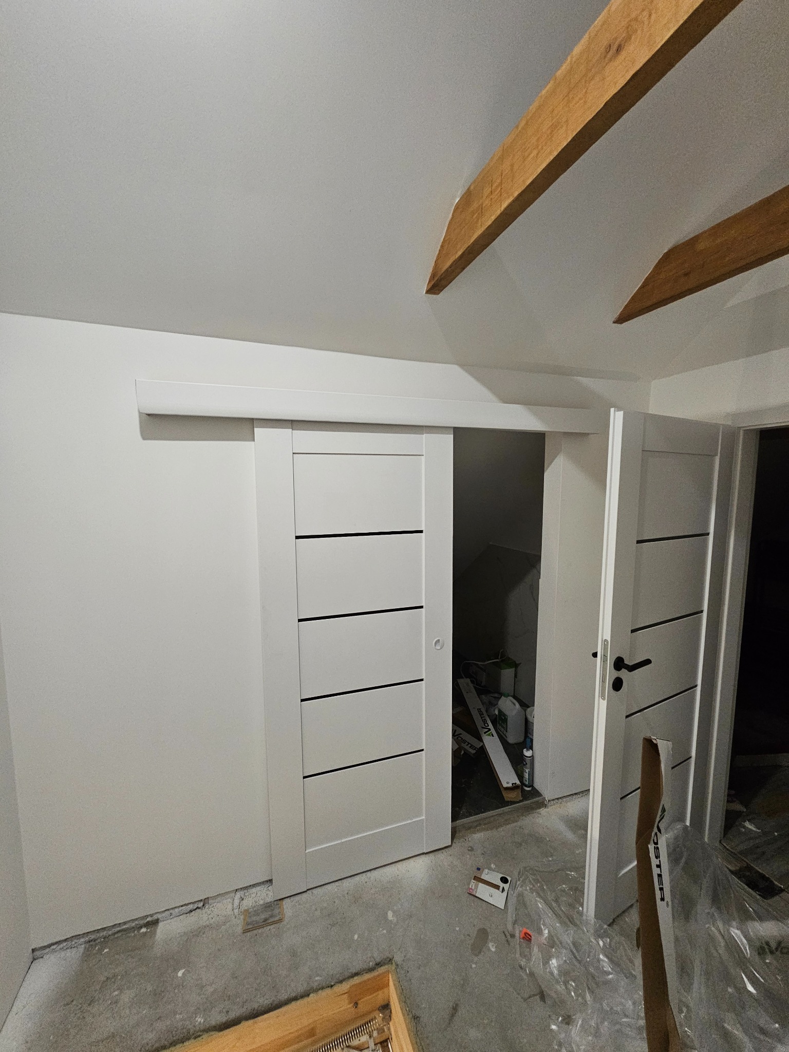 Białe, przesuwne drzwi z poziomymi frezami w nowym wnętrzu. Widoczne drewniane belki stropowe i surowa podłoga. W tle otwarte drzwi do pomieszczenia.