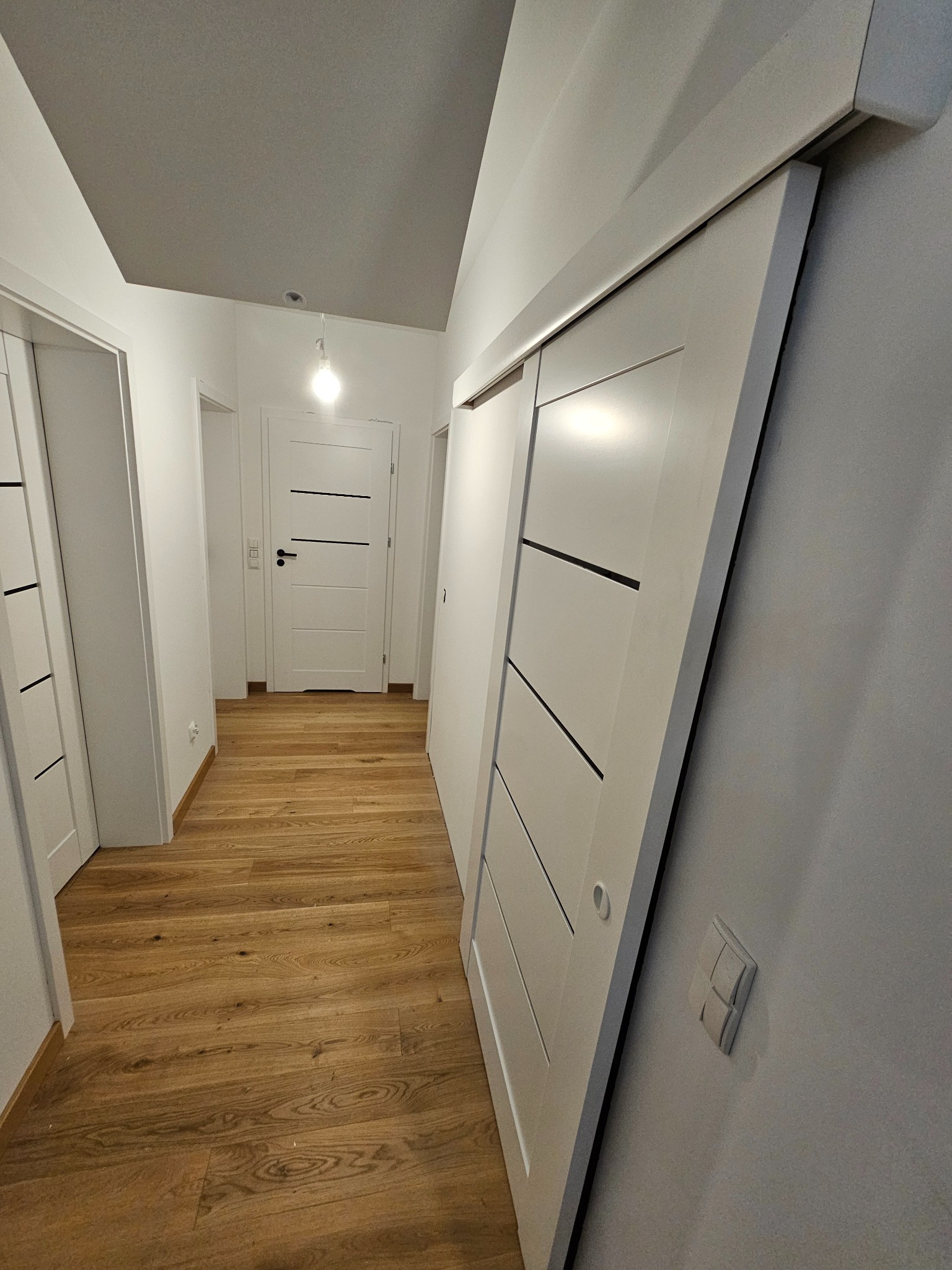 Białe drzwi przesuwne z czarnymi akcentami w jasnym korytarzu z drewnianą podłogą. Widoczne drzwi wejściowe w tle. Minimalistyczny design.