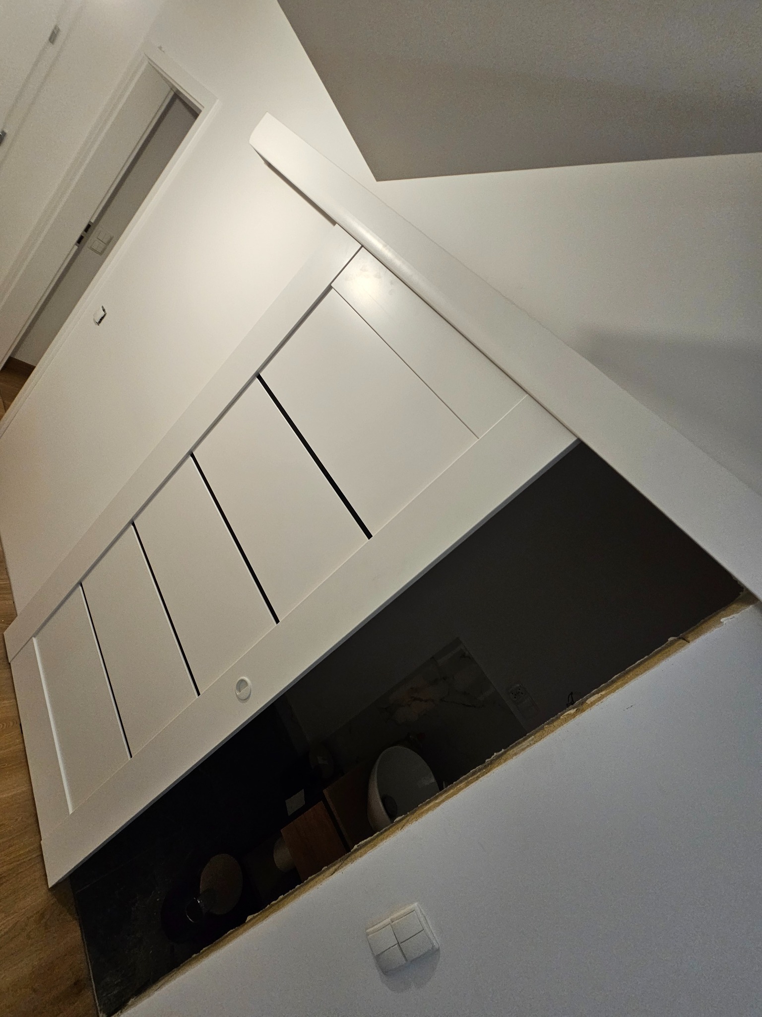 Biała zabudowa szafy wnękowej z czterema frontami, widoczna z boku, z otwartym otworem rewizyjnym. Widoczne elementy instalacji wewnątrz.