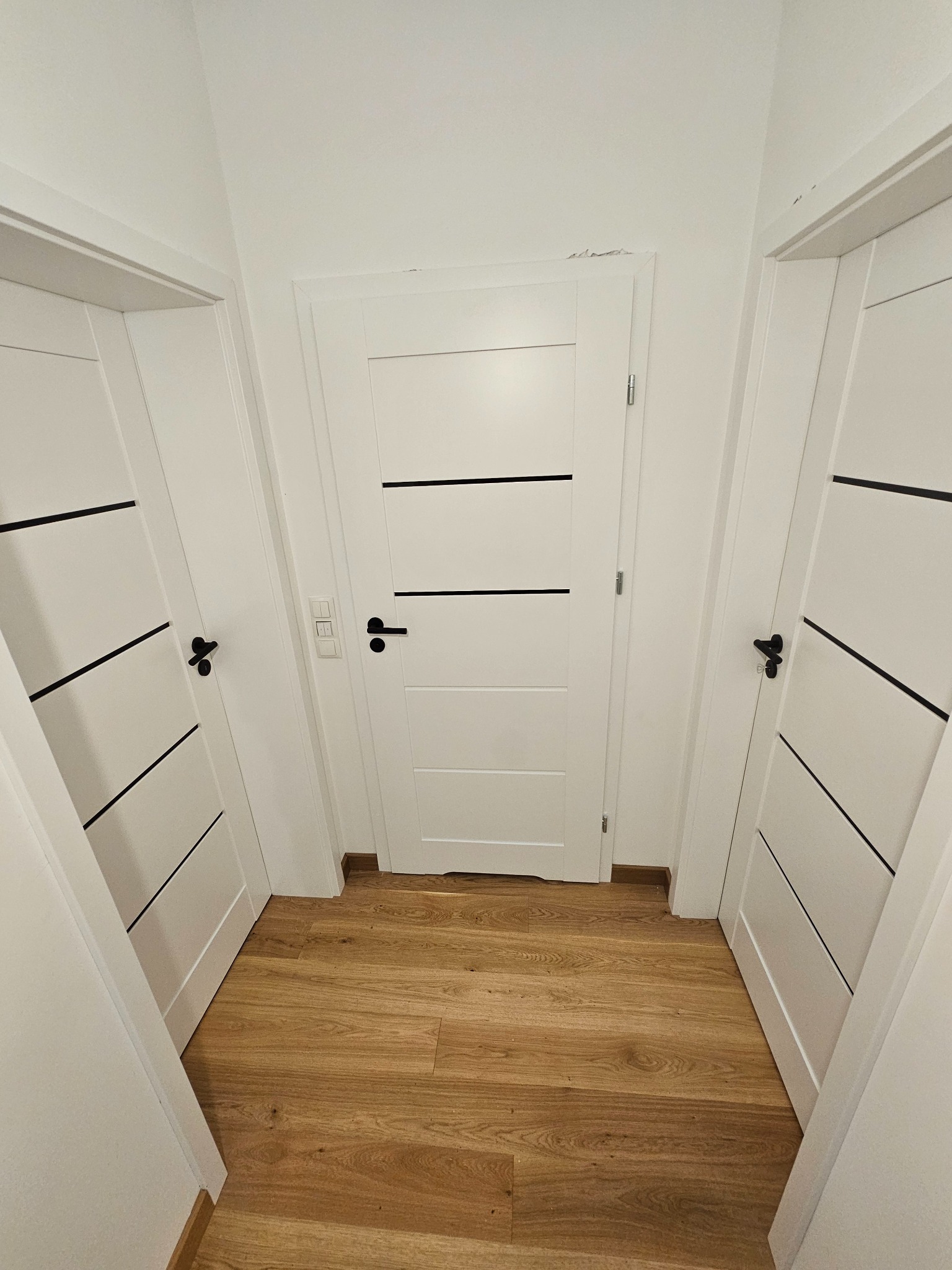 Białe, nowoczesne drzwi z czarnymi akcentami w korytarzu z drewnianą podłogą. Widoczne trzy skrzydła drzwiowe i włącznik światła.