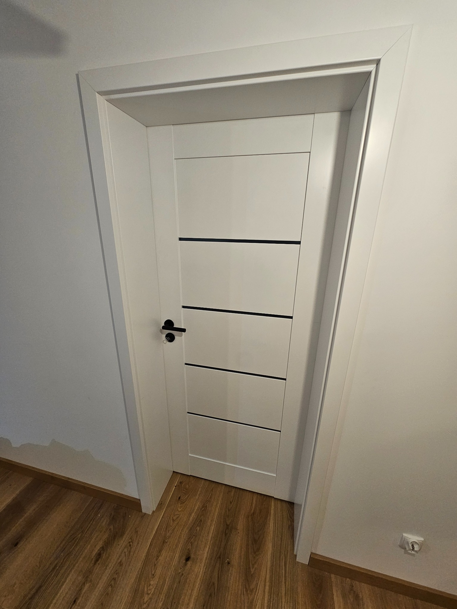 Nowoczesne, białe drzwi wewnętrzne z czarnymi, poziomymi akcentami i czarną klamką, wstawione w białą ościeżnicę, na tle ściany i drewnianej podłogi.