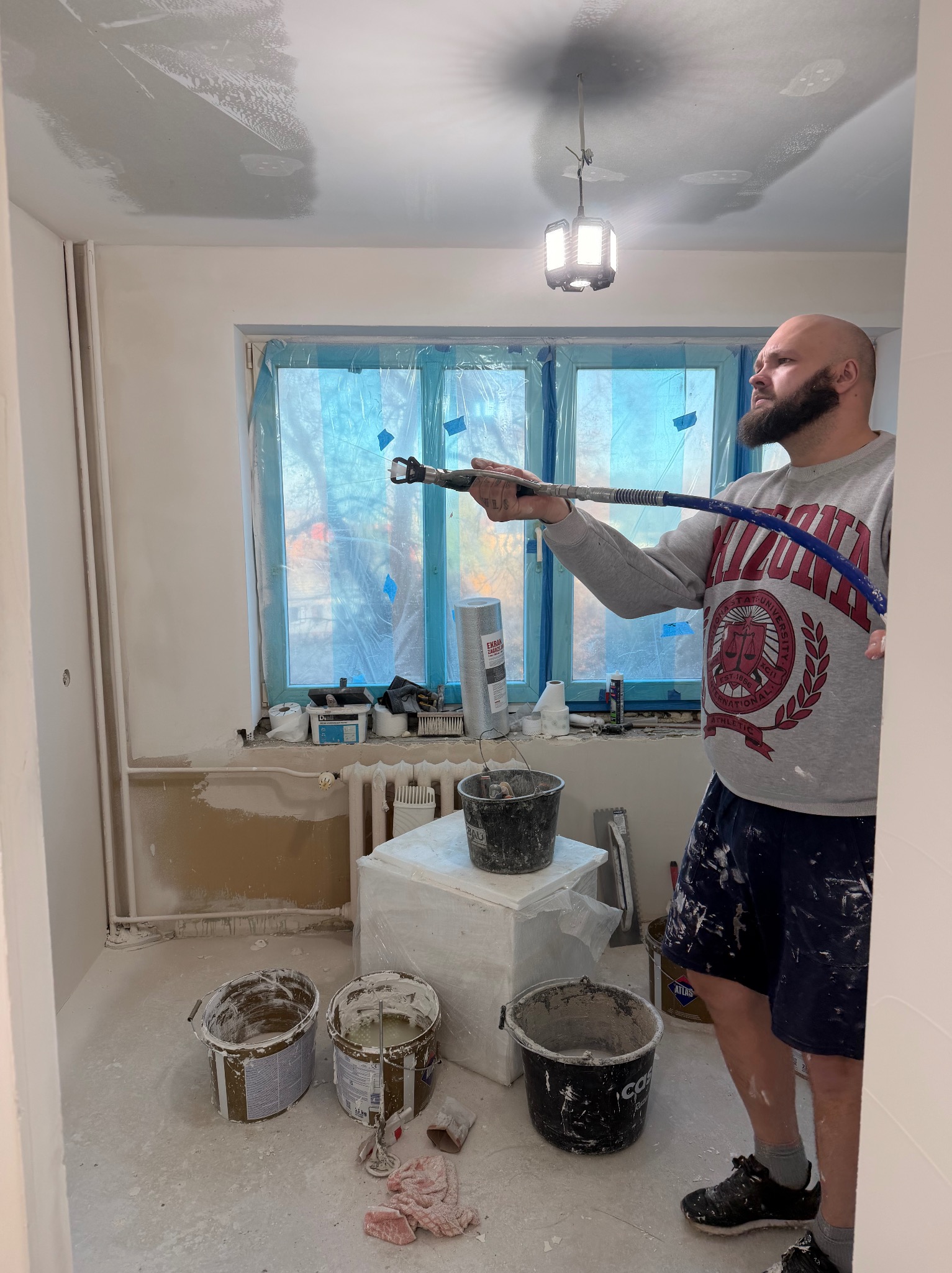 Aplikacja gładzi natryskowej w jasnym wnętrzu. Widoczny mężczyzna z pistoletem natryskowym, przygotowane wiadra z gładzią i zabezpieczone okno folią malarską.