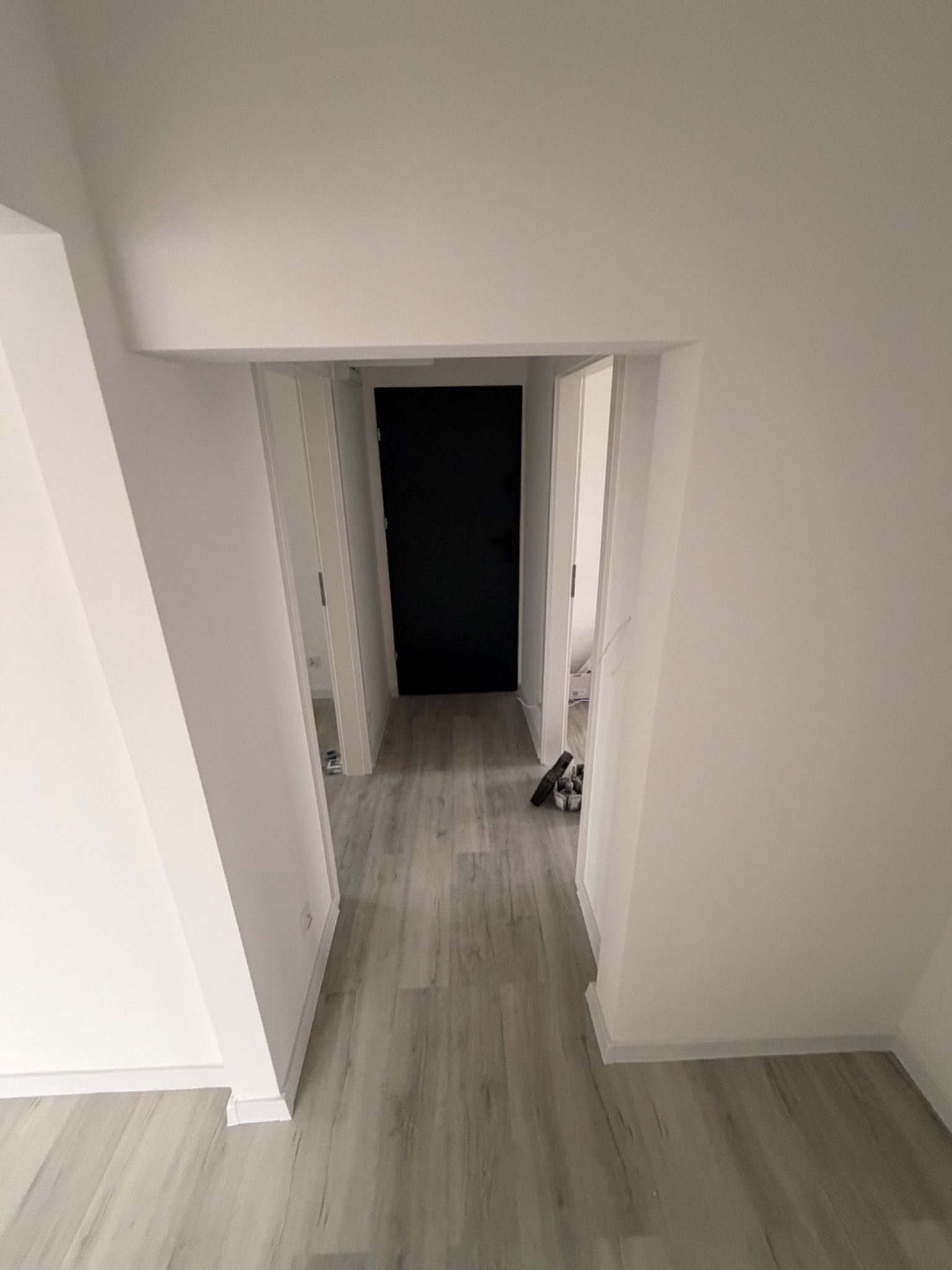 Wnętrze po remoncie: białe ściany, szara podłoga imitująca drewno, czarne drzwi w głębi. Widok z perspektywy wejścia, minimalistyczny design.