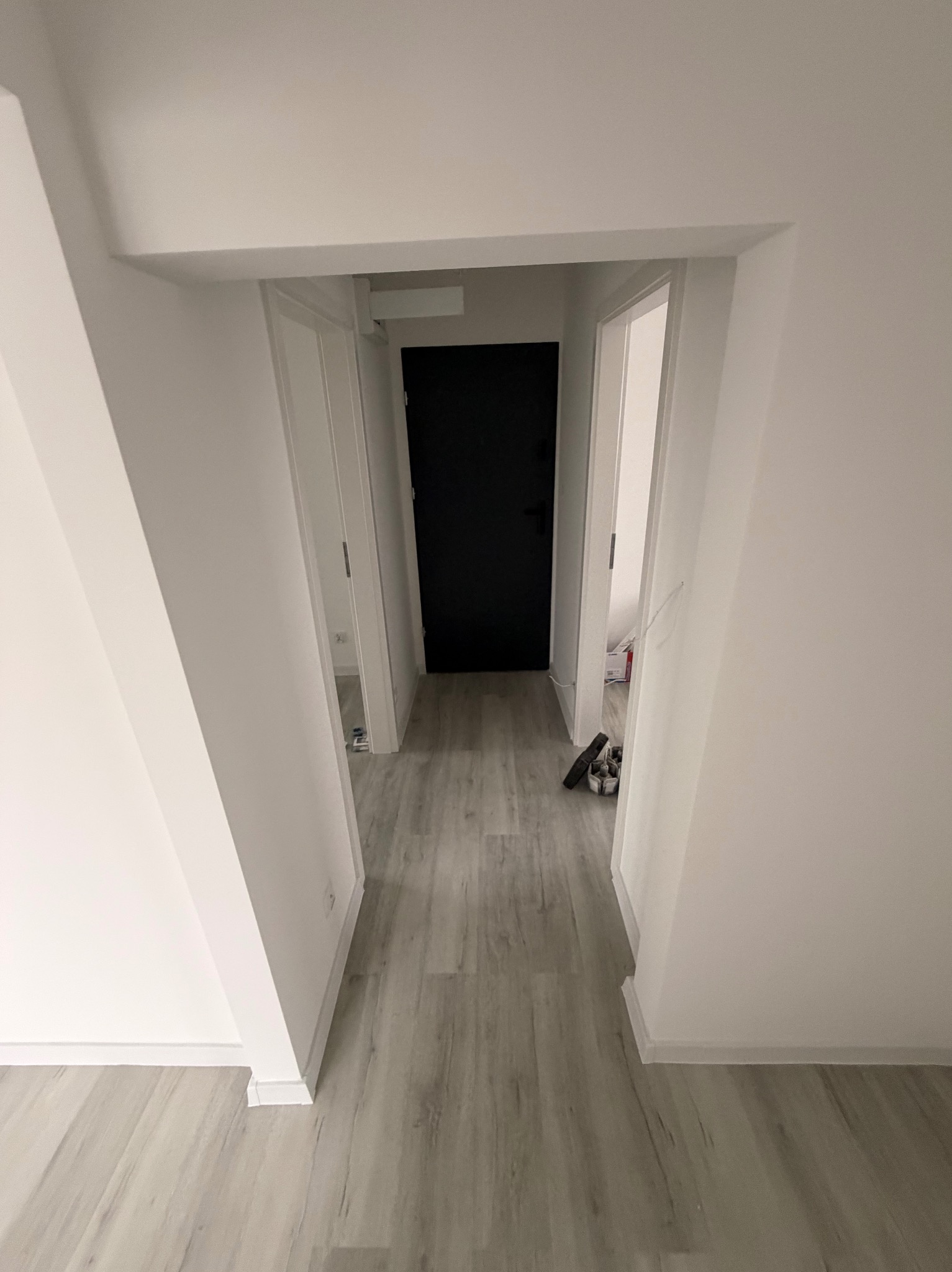 Białe wnętrze mieszkania z drewnopodobną podłogą, widok na korytarz z czarnymi drzwiami. Minimalistyczny design, świeżo po remoncie, widoczne listwy przypodłogowe.