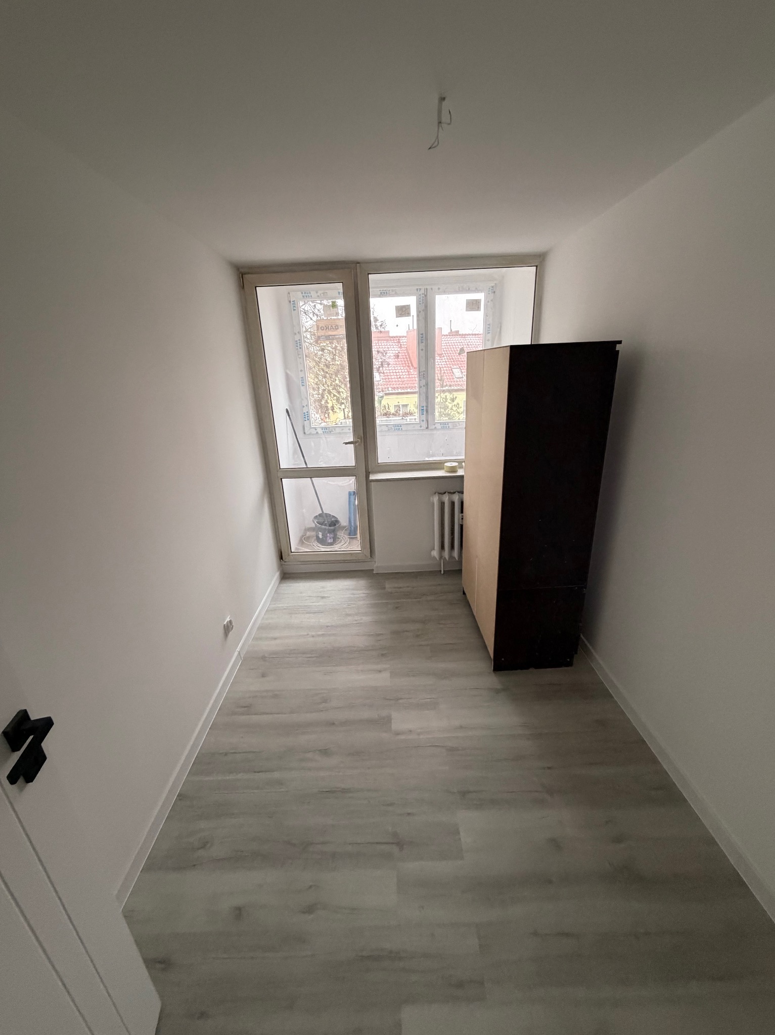 Pomalowane na biało wnętrze pokoju z panelami podłogowymi, widok na okno balkonowe z wiadrem i mopem, oraz ciemny mebel przy ścianie.