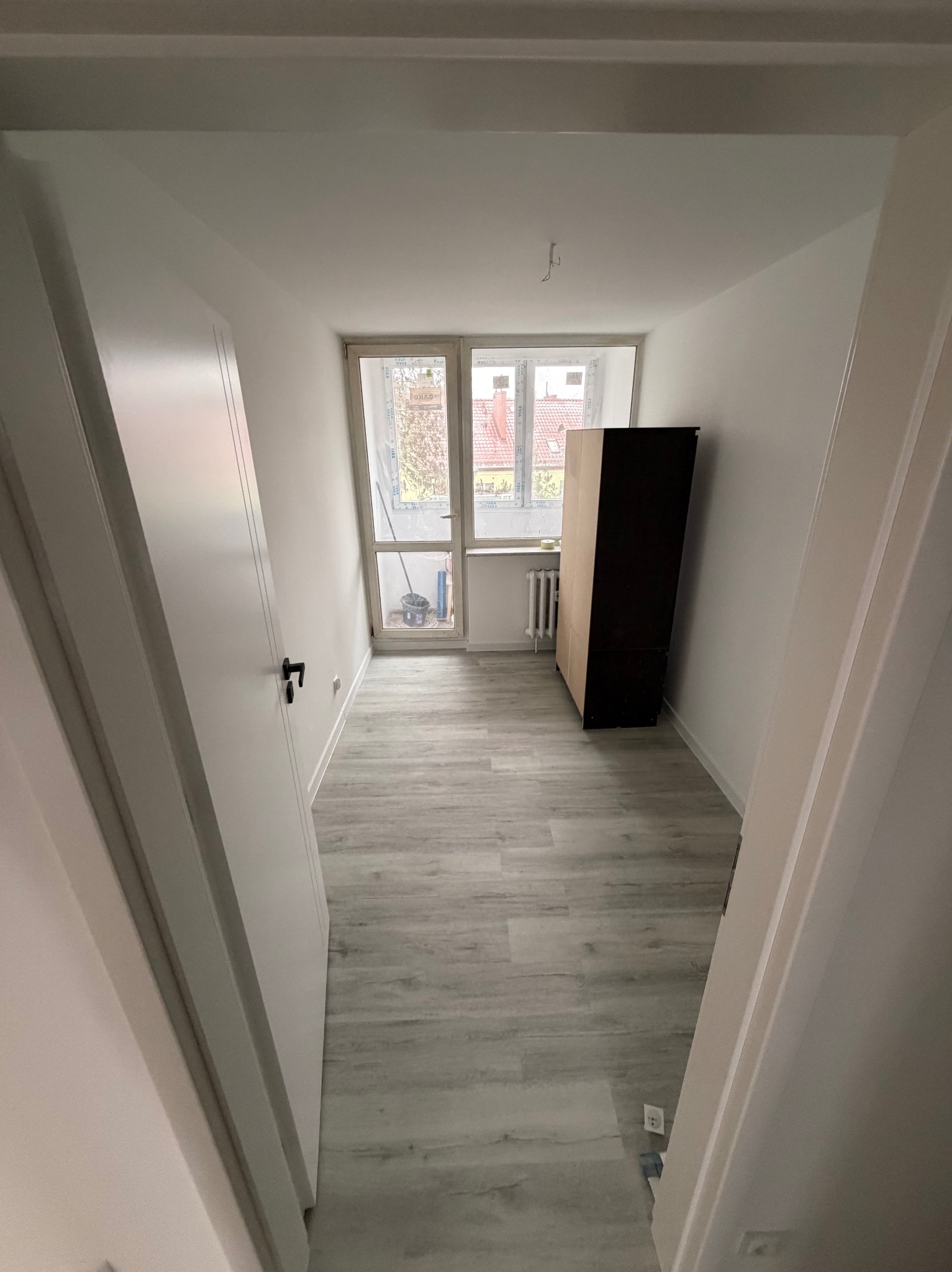 Jasne wnętrze po remoncie: szare panele, białe ściany i drzwi, okno z widokiem na zewnątrz. Minimalistyczny styl, czysty kadr, widoczne gniazdko elektryczne przy podłodze.