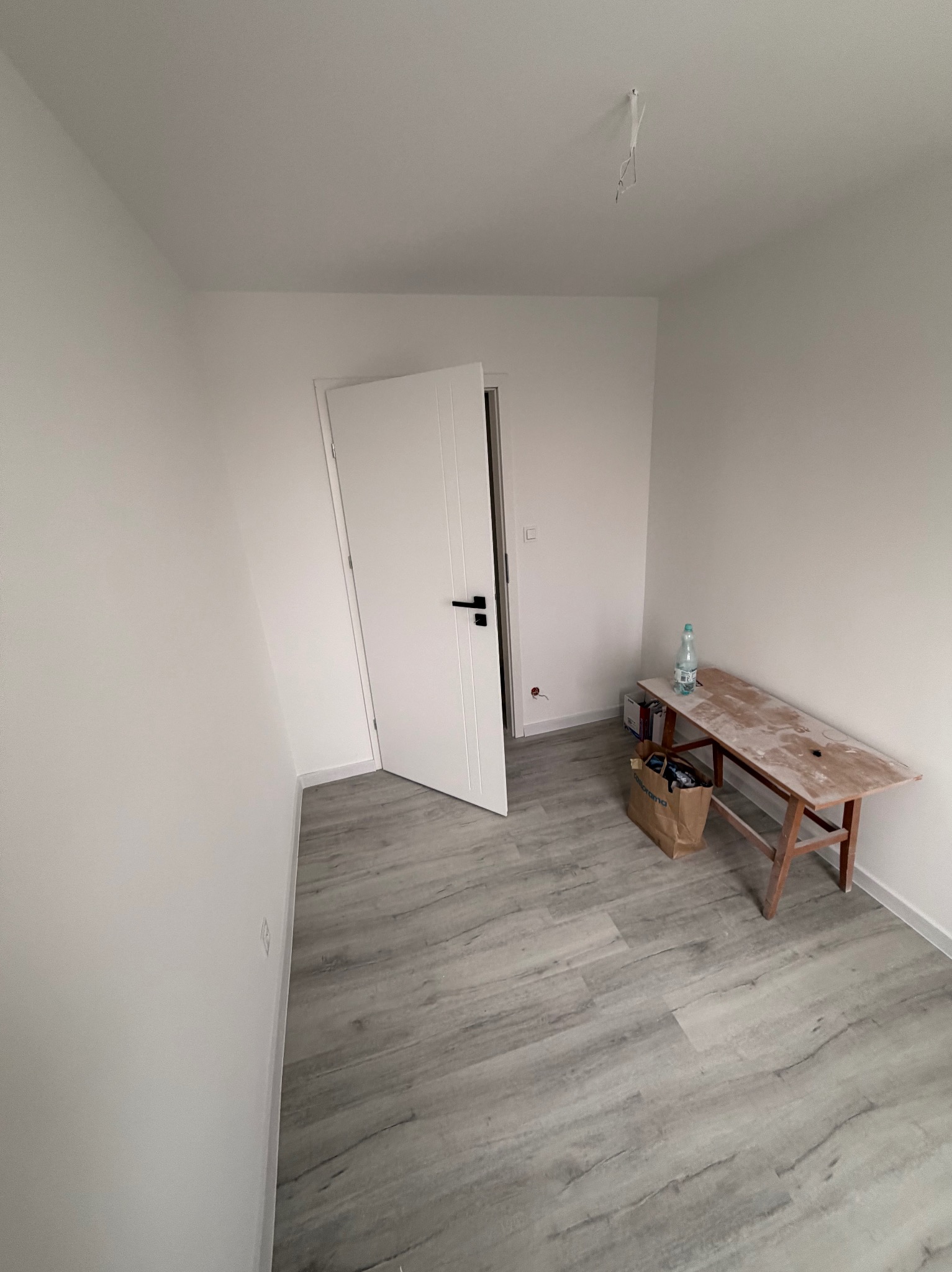 Wnętrze pokoju po remoncie: białe ściany, szara podłoga imitująca drewno, białe drzwi z czarną klamką, drewniana ławka i torba Castorama. Minimalistyczny wystrój.