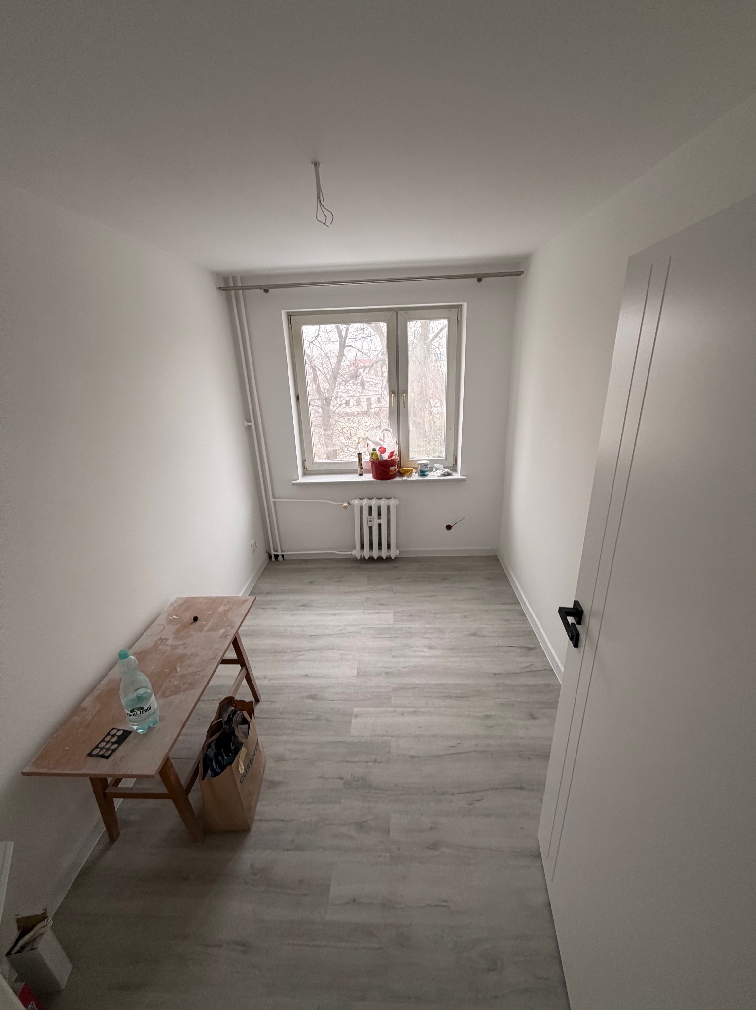 Wnętrze pokoju po malowaniu na biało, widoczne świeżo pomalowane ściany, okno z grzejnikiem i drewniana ławka z butelką wody. Podłoga wyłożona panelami.