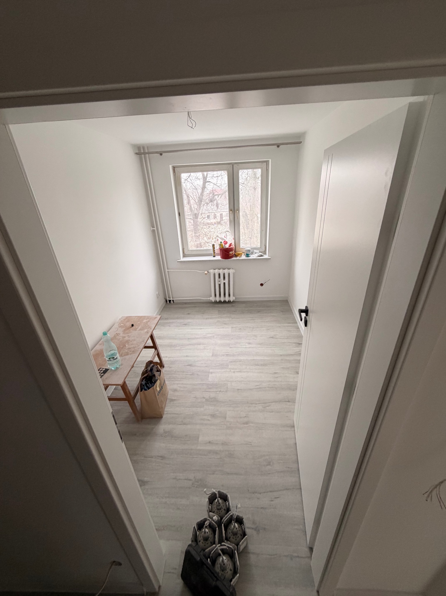 Jasne wnętrze po remoncie: białe ściany, panele podłogowe, okno z widokiem na drzewa. Widok zza futryny, narzędzia malarskie w rogu. Minimalistyczny styl.