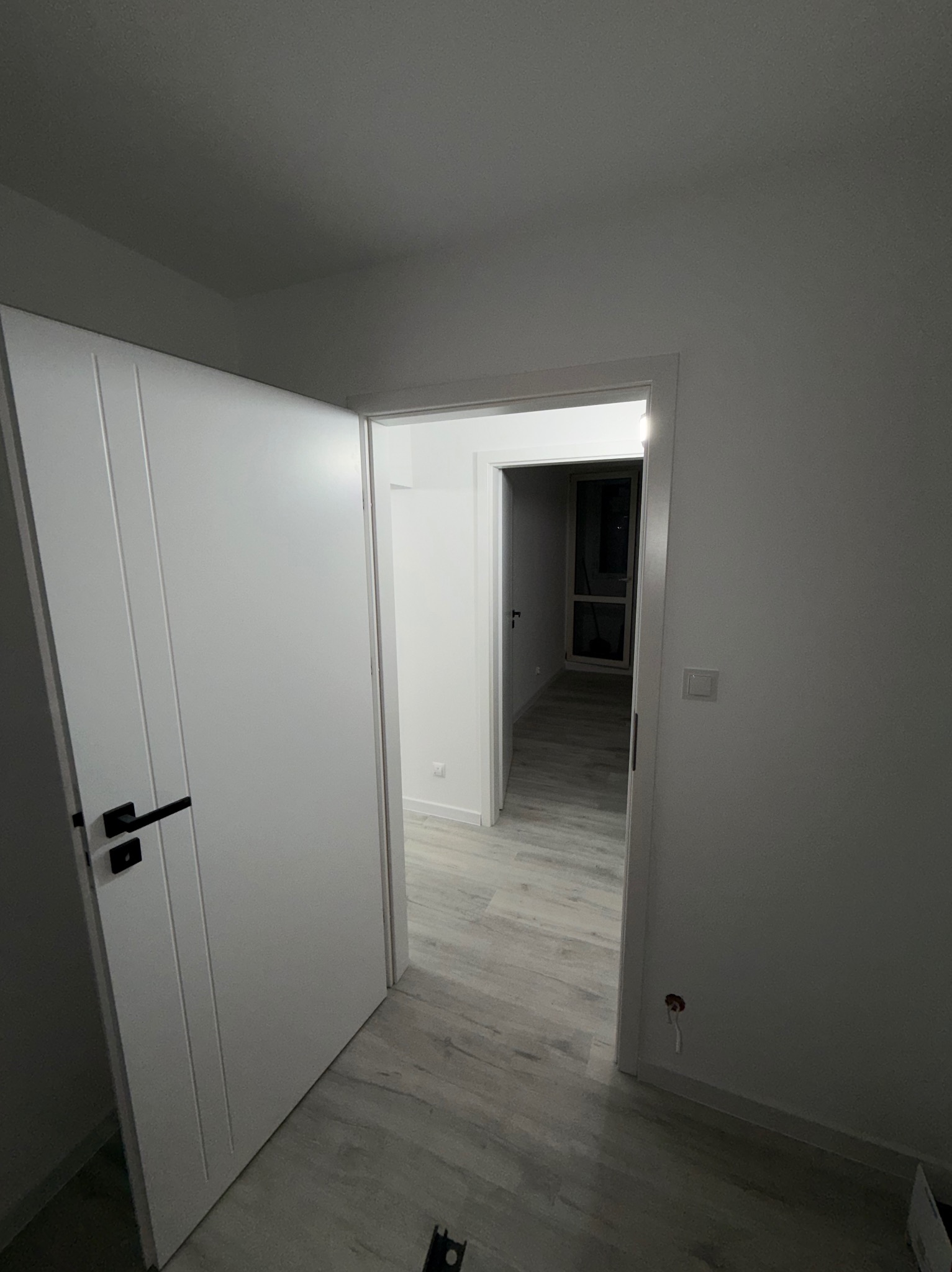 Nowoczesne, białe drzwi z czarną klamką, otwarte do wnętrza wykończonego mieszkania z panelami podłogowymi i widokiem na kolejne pomieszczenia. Minimalistyczny design.