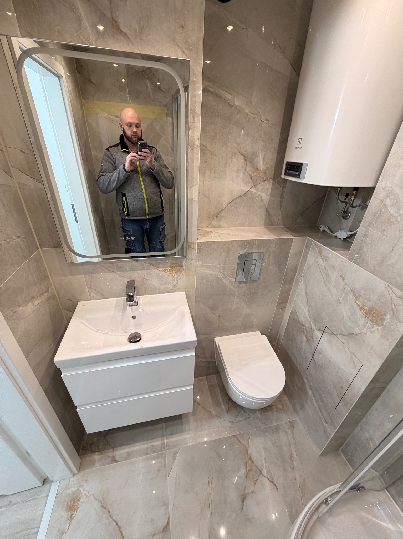 Wykończona łazienka w beżowych płytkach marmuropodobnych. Widoczna umywalka z szafką, toaleta, bojler i lustro, w którym odbija się osoba robiąca zdjęcie.