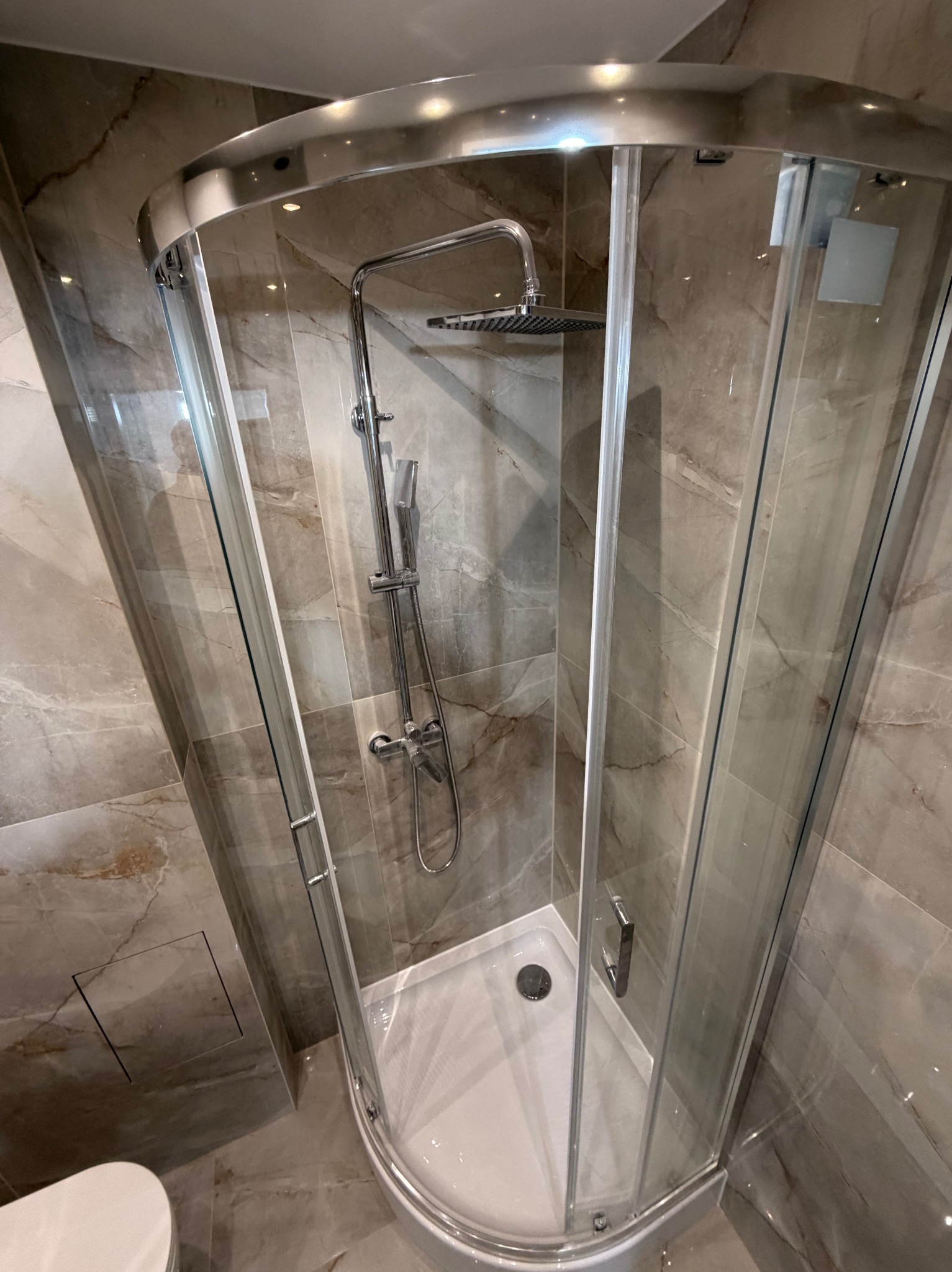 Nowoczesna kabina prysznicowa z chromowanymi elementami i deszczownicą, wykończona płytkami imitującymi marmur. Widoczny fragment toalety. Górny kadr.