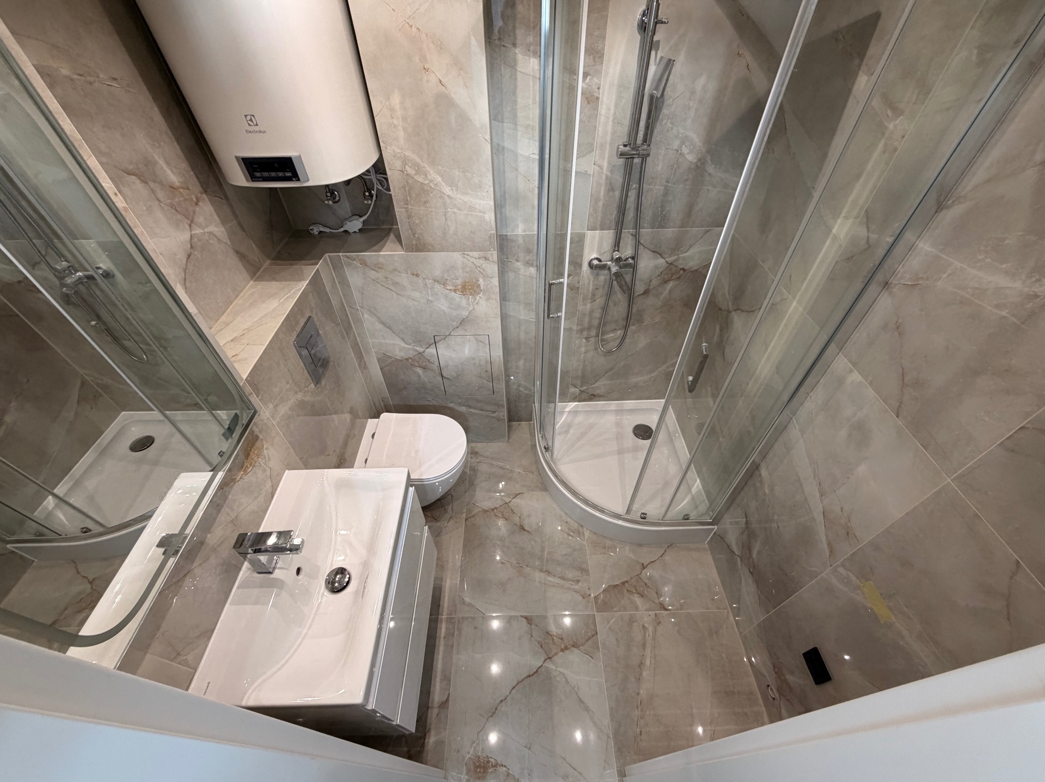 Wykończona łazienka z kabiną prysznicową, umywalką i toaletą, wyłożona płytkami imitującymi marmur. Widoczny bojler elektryczny. Ujęcie z góry, podkreślające przestrzeń.