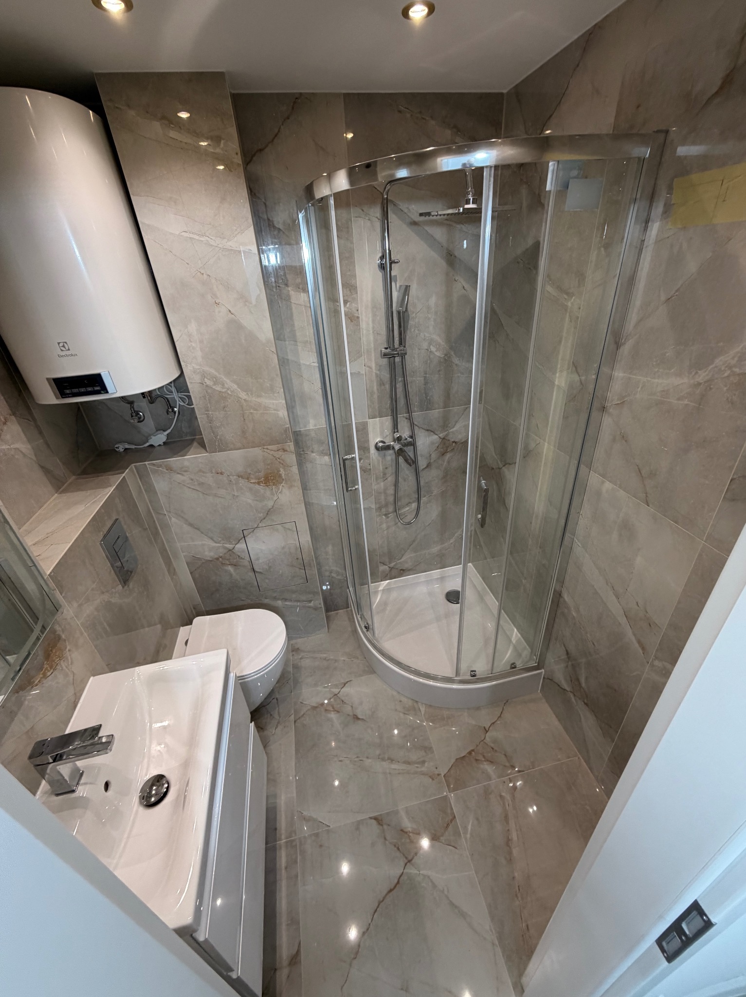 Wykończona łazienka z kabiną prysznicową, umywalką i toaletą. Płytki imitujące marmur na ścianach i podłodze. Widoczny bojler elektryczny. Nowoczesny design.