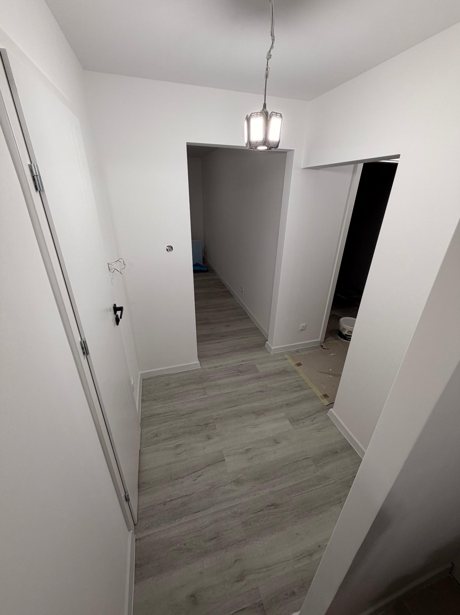 Białe wnętrze po remoncie: korytarz z szarą podłogą imitującą drewno, białe ściany i drzwi, widoczna lampa sufitowa i fragment puszki z farbą.