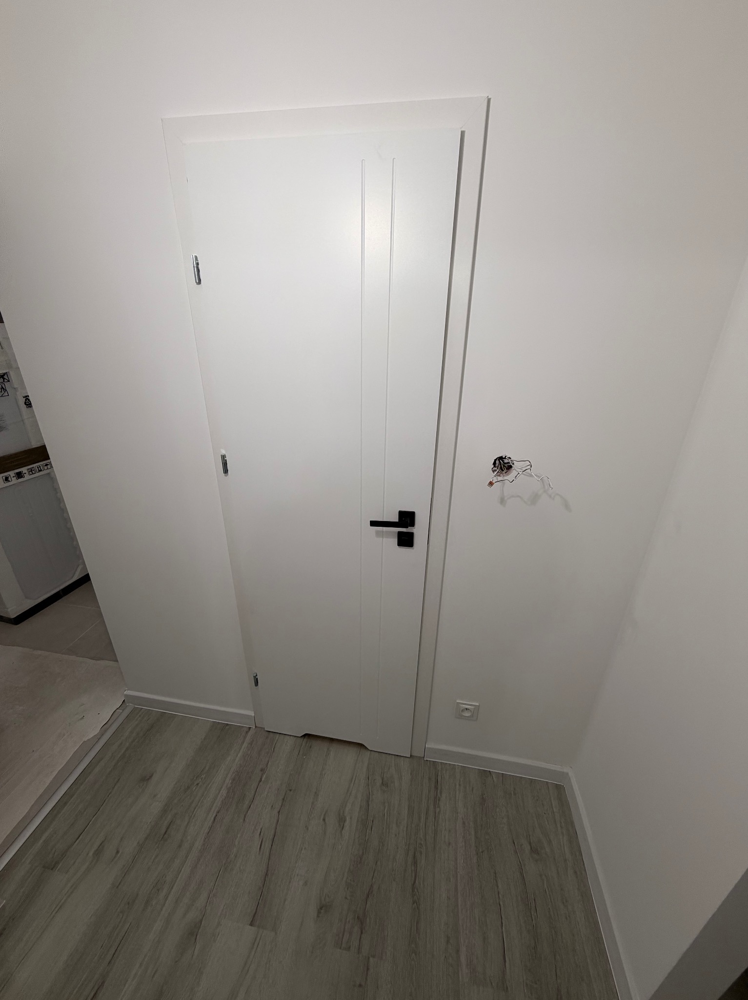 Białe drzwi z czarną klamką w białym pomieszczeniu z szarą podłogą. Widoczne wystające kable elektryczne i gniazdko. Minimalistyczny design.