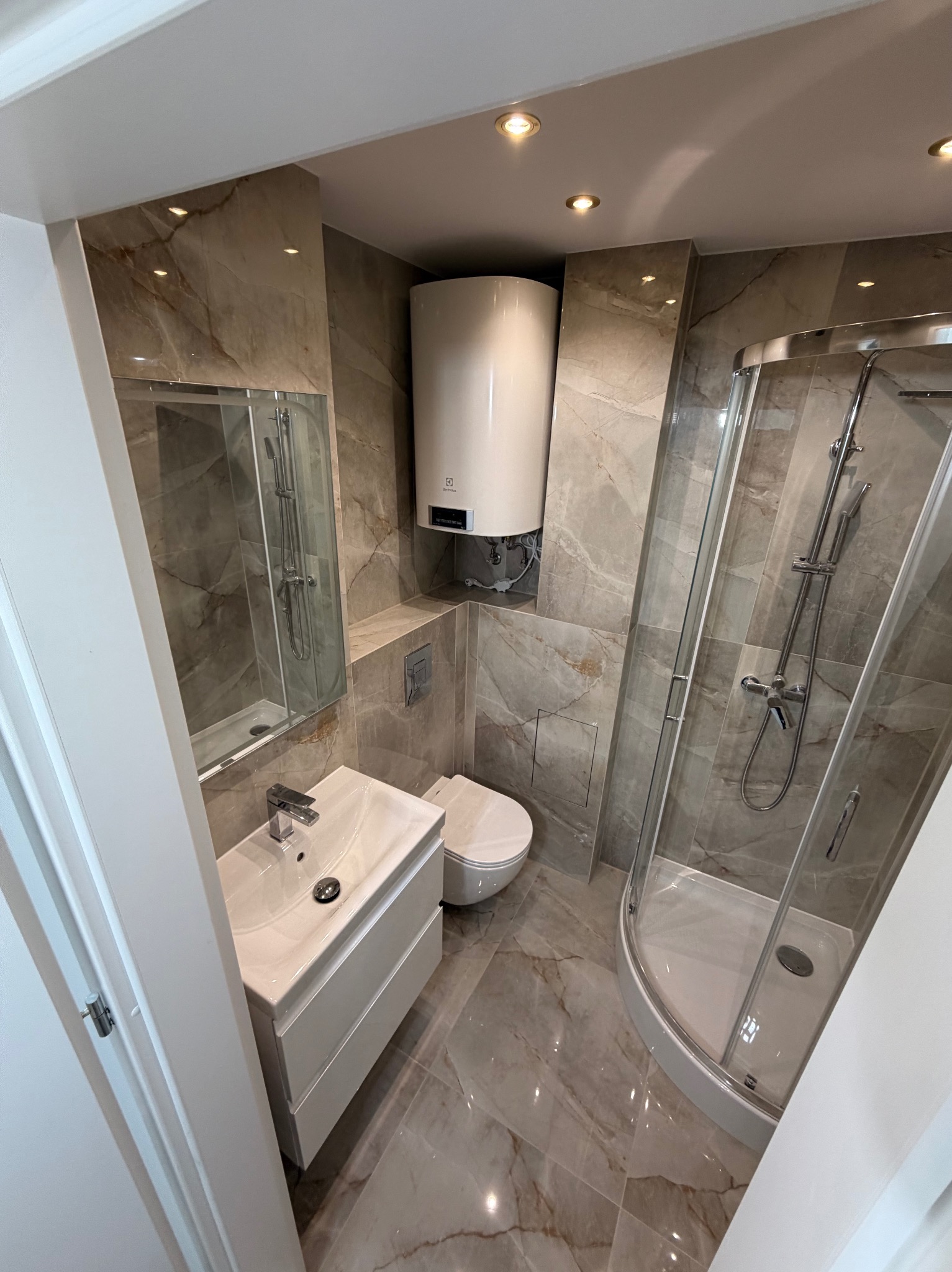 Wykończona łazienka w beżowych płytkach imitujących marmur. Kabina prysznicowa, umywalka z szafką, toaleta i bojler elektryczny. Oświetlenie punktowe.