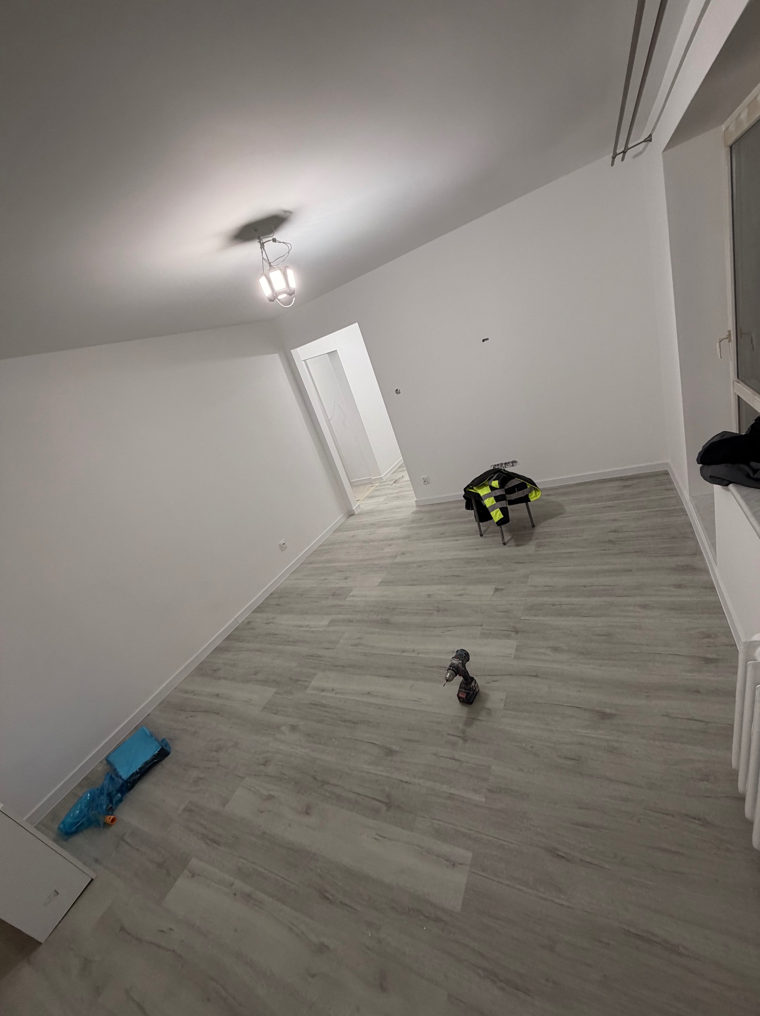 Wnętrze po remoncie: świeżo pomalowane ściany na biało, jasna podłoga, narzędzia pracy (wiertarka) i odzież robocza. Widok na odnowione pomieszczenie z nowym oświetleniem.