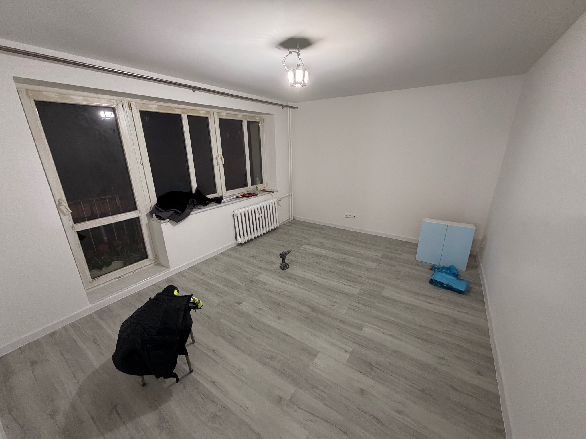 Pusty pokój po remoncie z nową podłogą, grzejnikiem i oknami. W tle narzędzie - wiertarka. Na krześle leży kurtka robocza. Widoczne materiały budowlane.