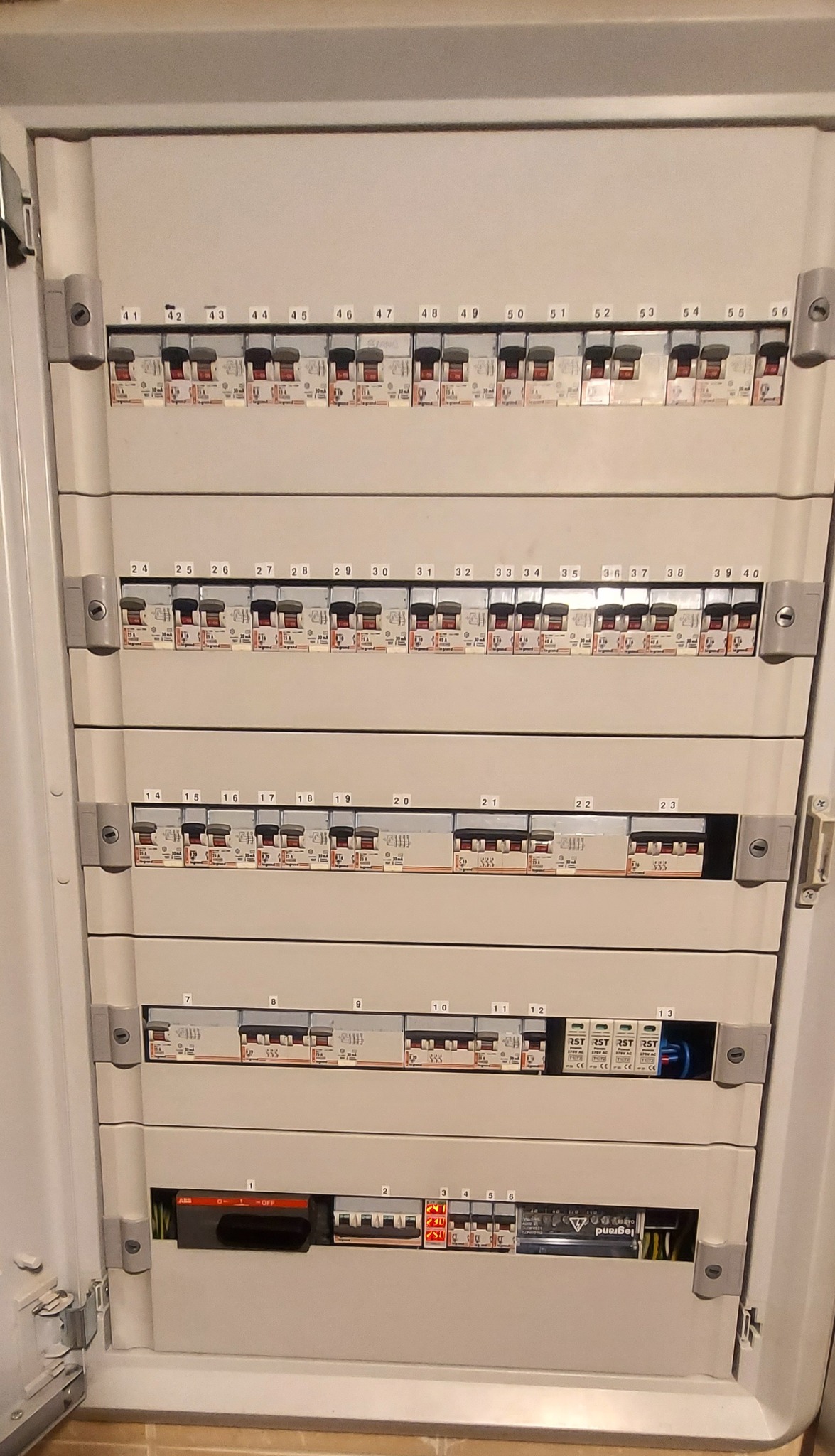 Otwarta skrzynka rozdzielcza z bezpiecznikami i wyłącznikami różnicowoprądowymi, widoczne numerowane moduły na szynach DIN, niektóre w pozycji 'ON', inne 'OFF', z przewodami w tle.