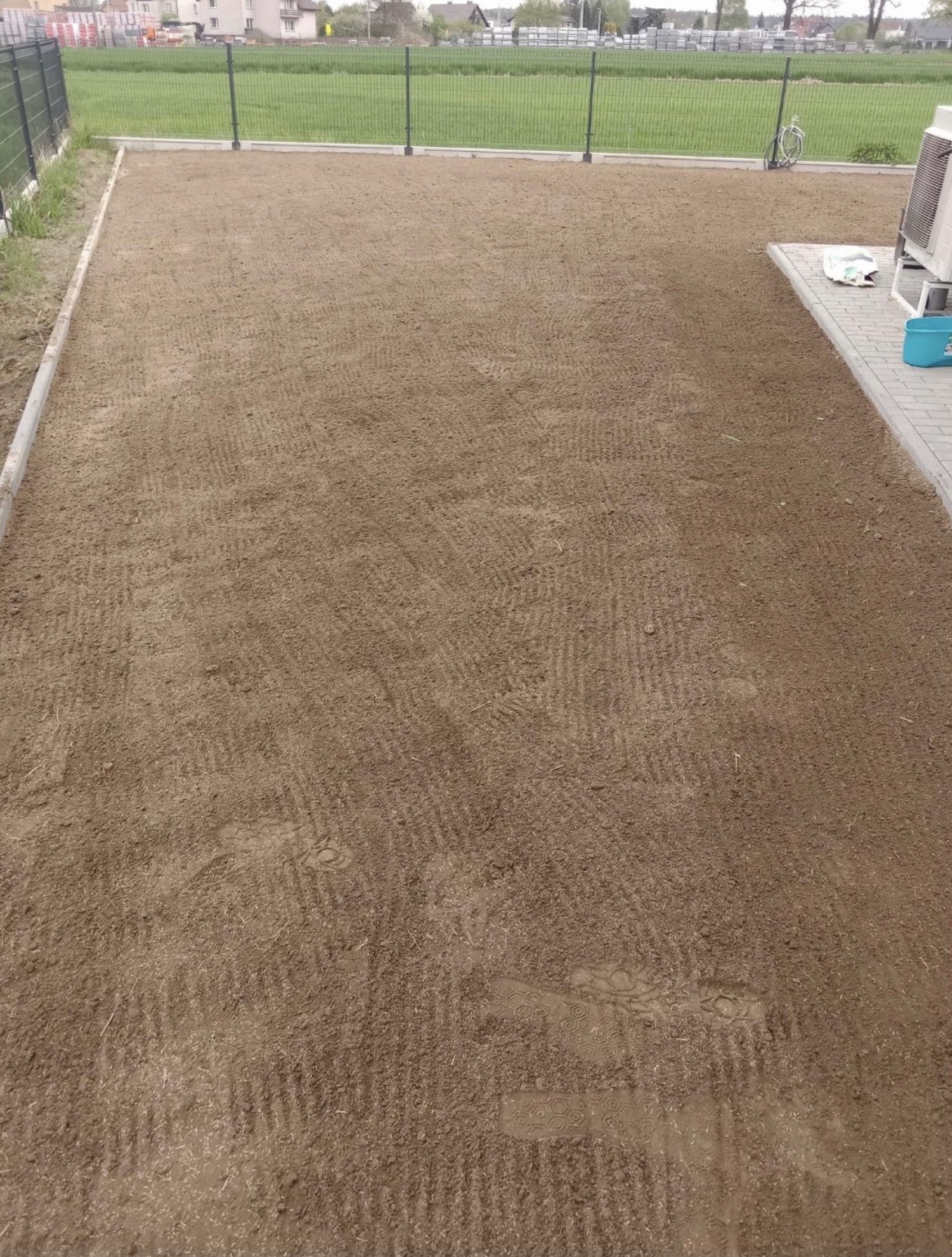 Przygotowany teren pod trawnik, wyrównana ziemia z widocznymi śladami po bronowaniu, obrzeża z kostki, w tle ogrodzenie panelowe i zabudowa jednorodzinna.
