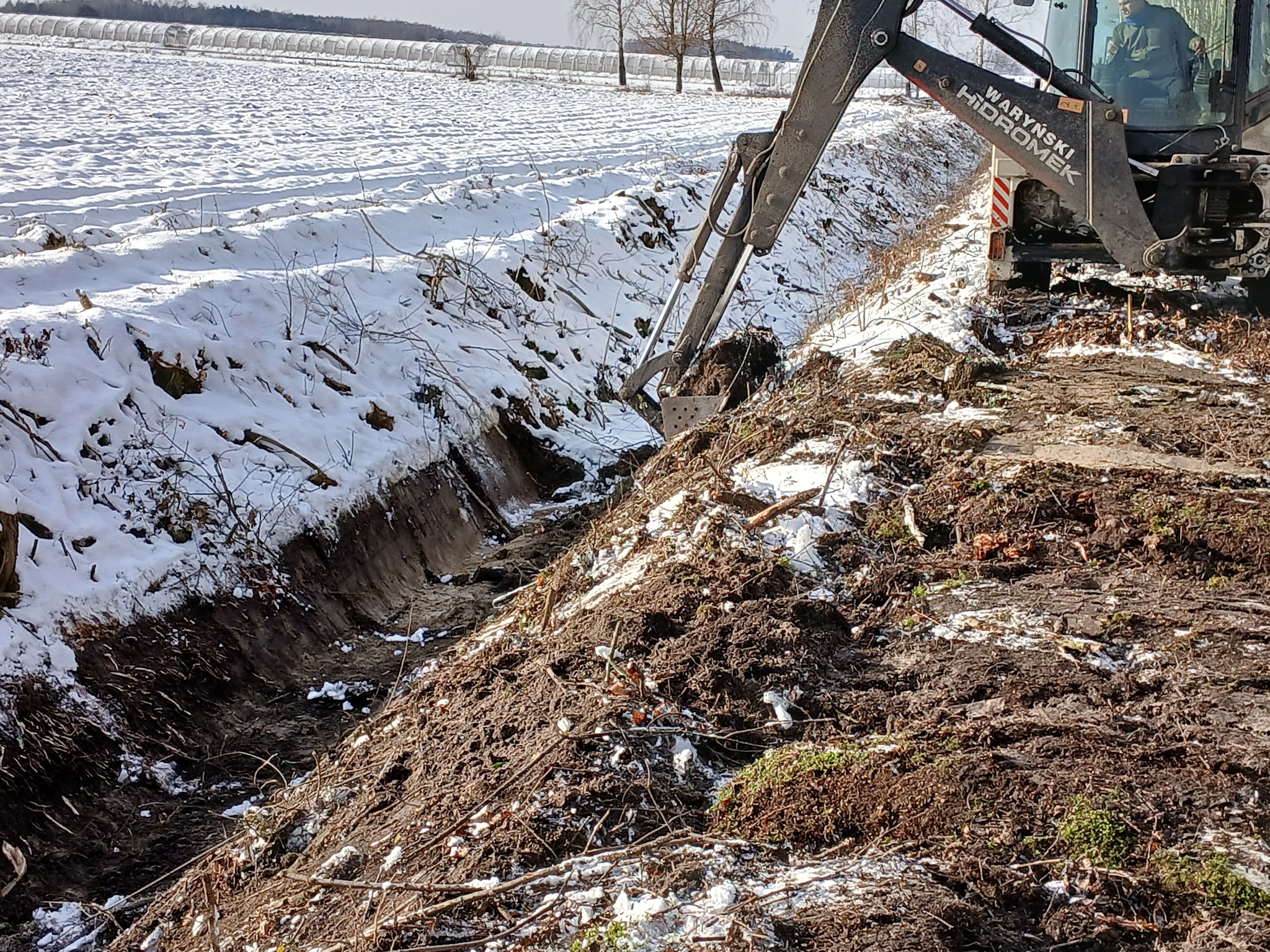 Koparka wykonuje wykop w ziemi pokrytej śniegiem, widoczne ślady opon i operator w kabinie.