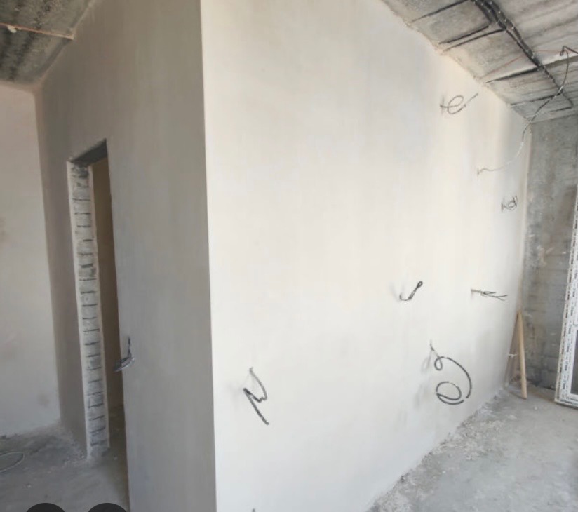 Świeżo położone, gładkie tynki maszynowe na ścianach w surowym wnętrzu. Widoczne wystające kable elektryczne i surowy betonowy sufit. W tle ościeżnica drzwi.