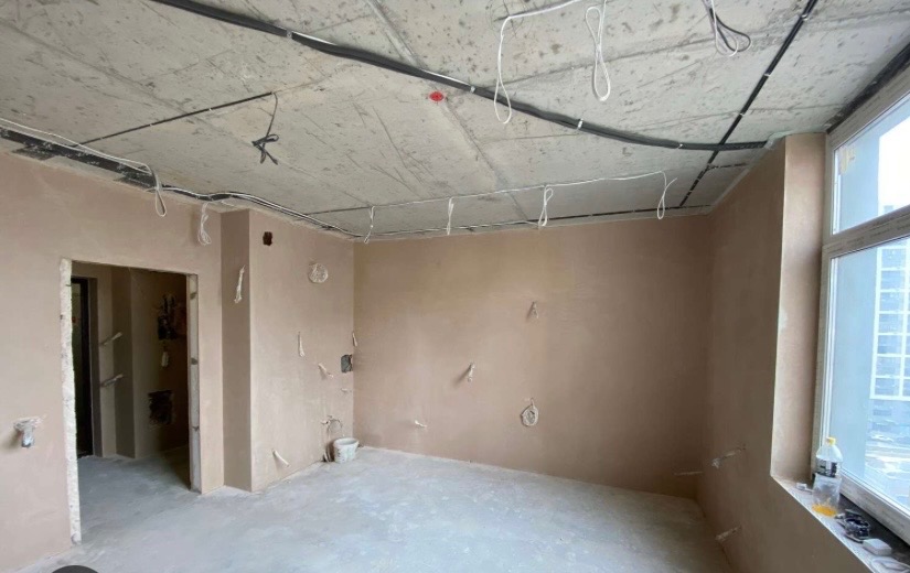 Wnętrze po nałożeniu gładzi: ściany pokryte jasnym tynkiem, widoczne przewody elektryczne pod sufitem, okno z widokiem na miasto. Minimalistyczny, surowy stan wykończenia.