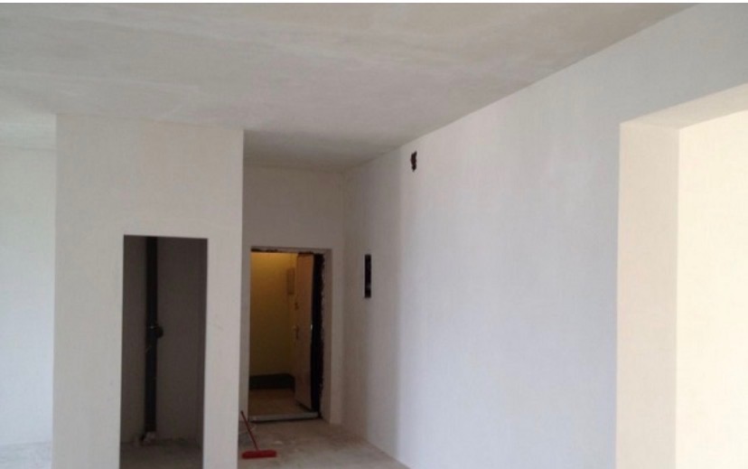 Wnętrze po gładziowaniu: białe ściany, sufit, otwory drzwiowe. Widoczne ślady po pracach wykończeniowych, czerwona zmiotka w rogu. Minimalistyczny efekt.