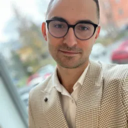 Portret mężczyzny w okularach i beżowej marynarce w drobną kratę, stojącego na tle okna z widokiem na budynek i samochody.